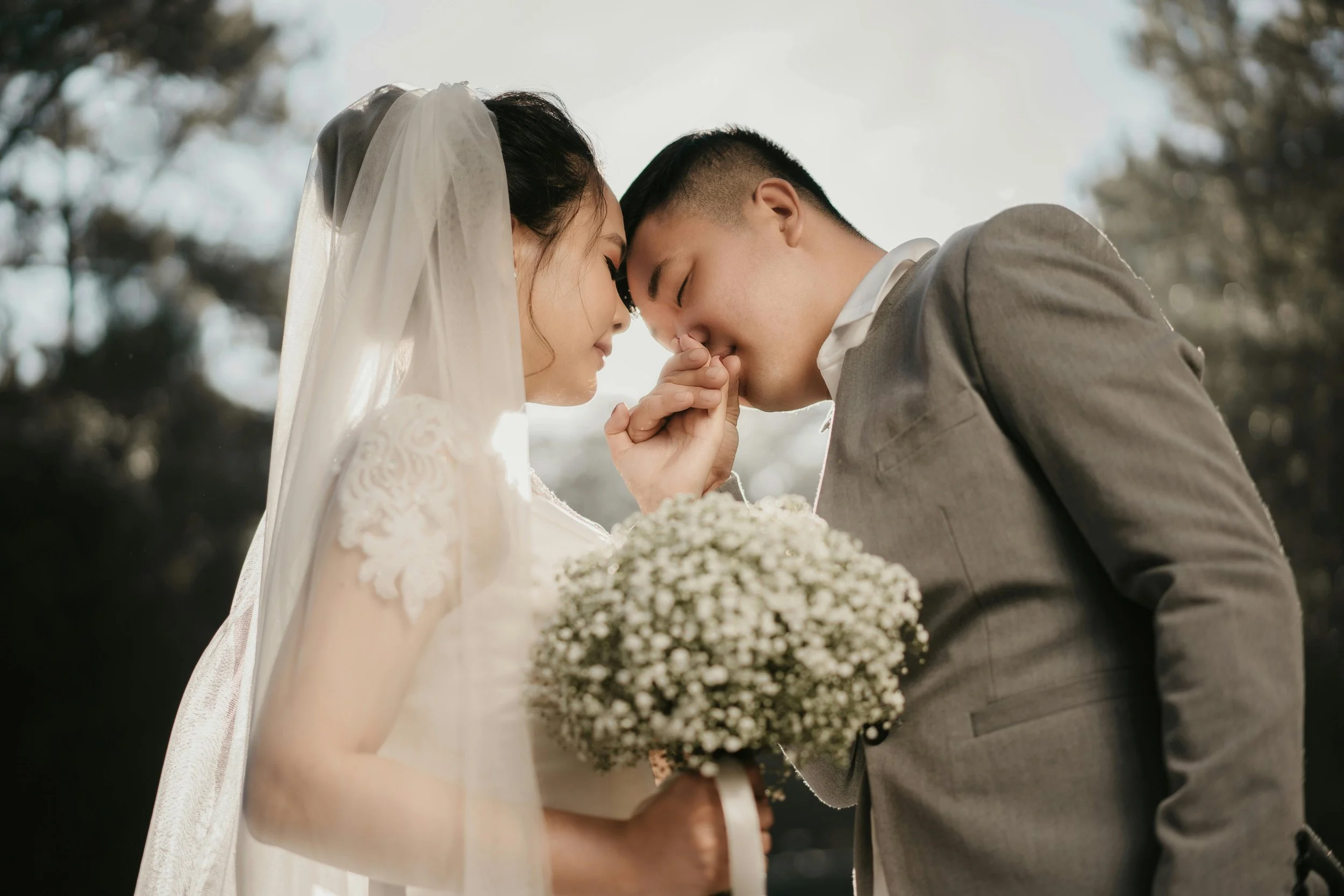 A bride and groom with foreheads touching and eyes closed, smiling, during their wedding ceremony outside. The bride is wearing a white lace wedding dress and veil, holding a bouquet of white flowers. The groom is dressed in a gray suit with a white shirt. They are touching hands in a tender moment, with a blurred outdoor background of trees.