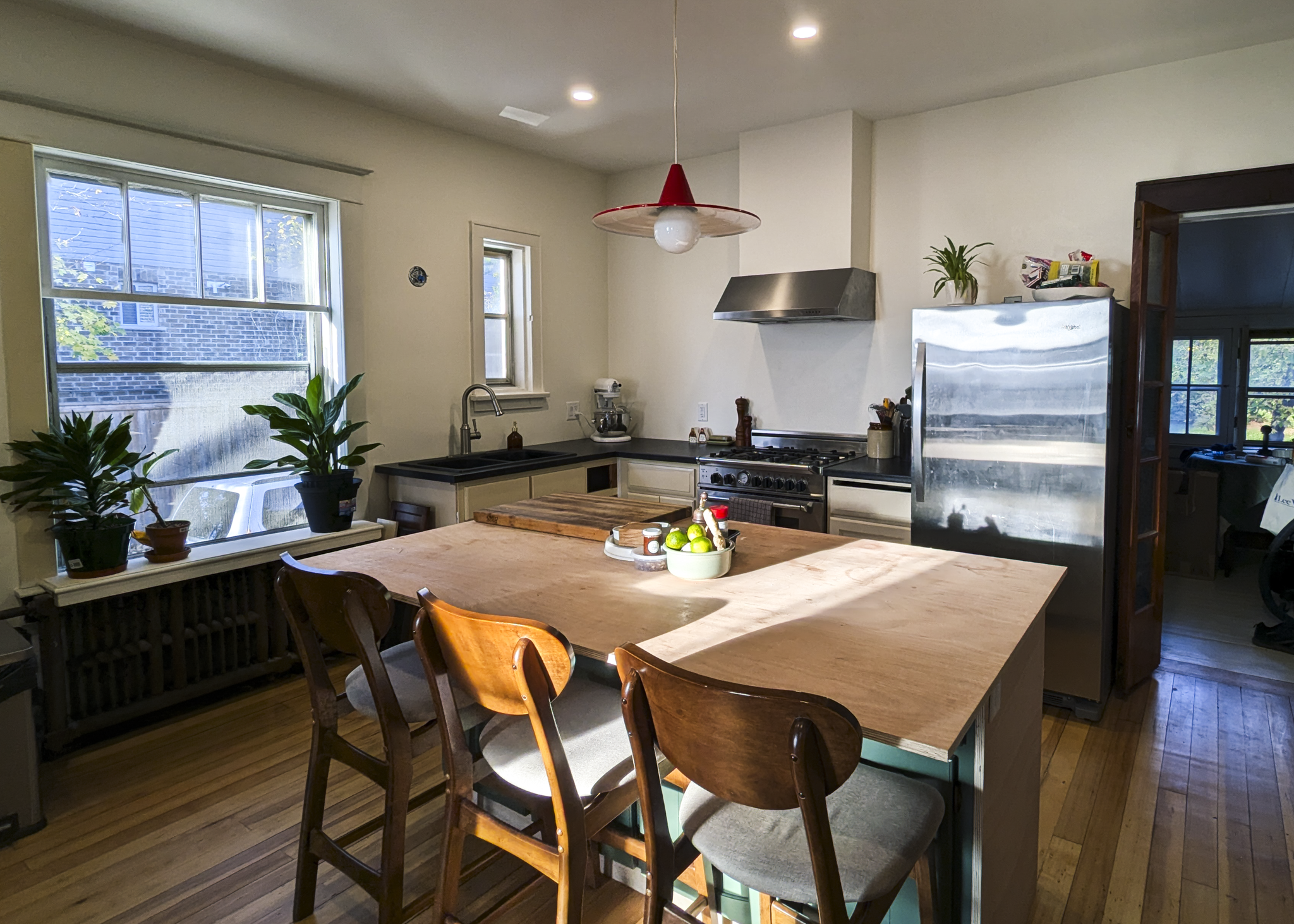 A kitchen with a wooden table, surrounded by wooden chairs. There are potted plants on the window sill, a sink under the window, and a stove with a stainless steel range hood. A refrigerator is on the right, with some items on top, and there's a doorway leading to another room. The lighting includes recessed ceiling lights and a red and transparent pendant lamp.