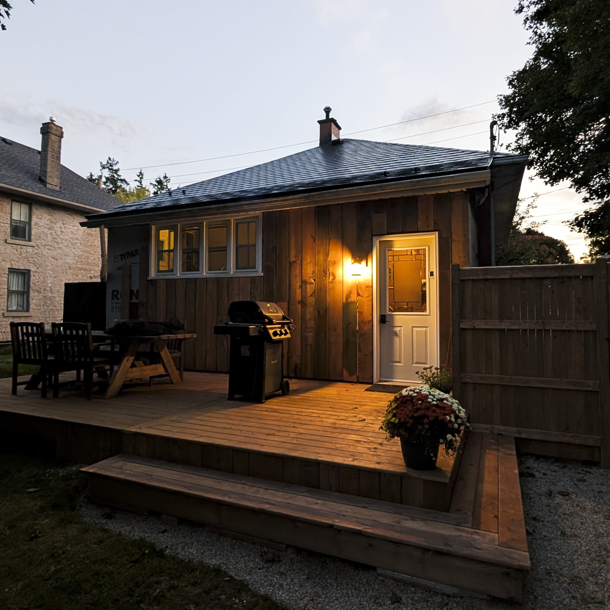 A house with a wooden exterior and a porch with a glowing light inside. There are two chairs and a picnic table on the porch. A grill is on the deck, and there is a flower arrangement on the ground near the porch. The house has a sloped roof with a chimney, and the evening sky is visible in the background.