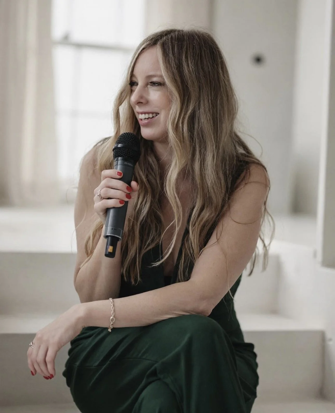 A woman with long wavy blonde hair holding a microphone, sitting on stairs near a window.