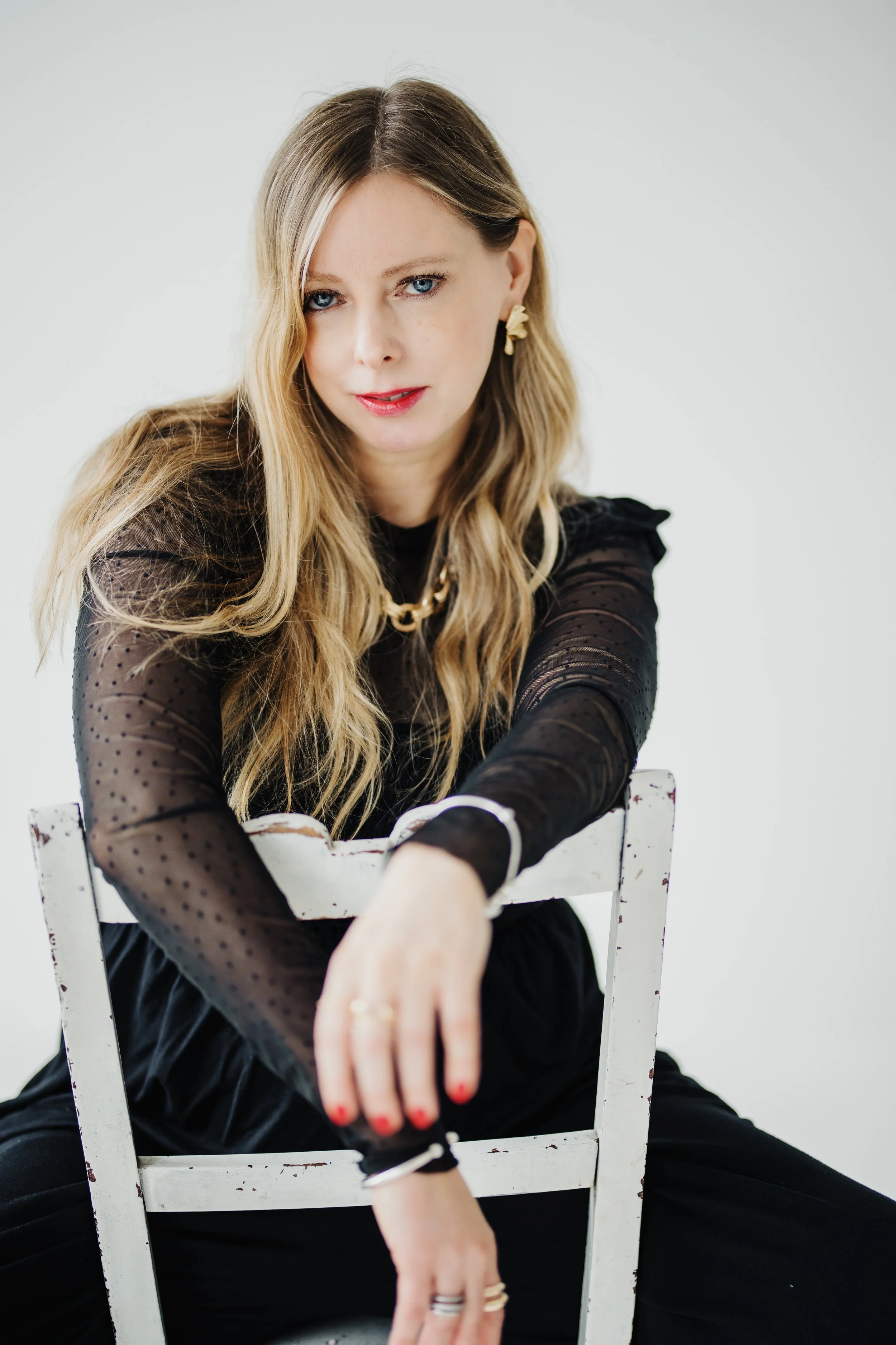 A woman with long, wavy blonde hair wearing a black sheer polka dot top, sitting on a white wooden chair, looking at the camera with a confident expression.