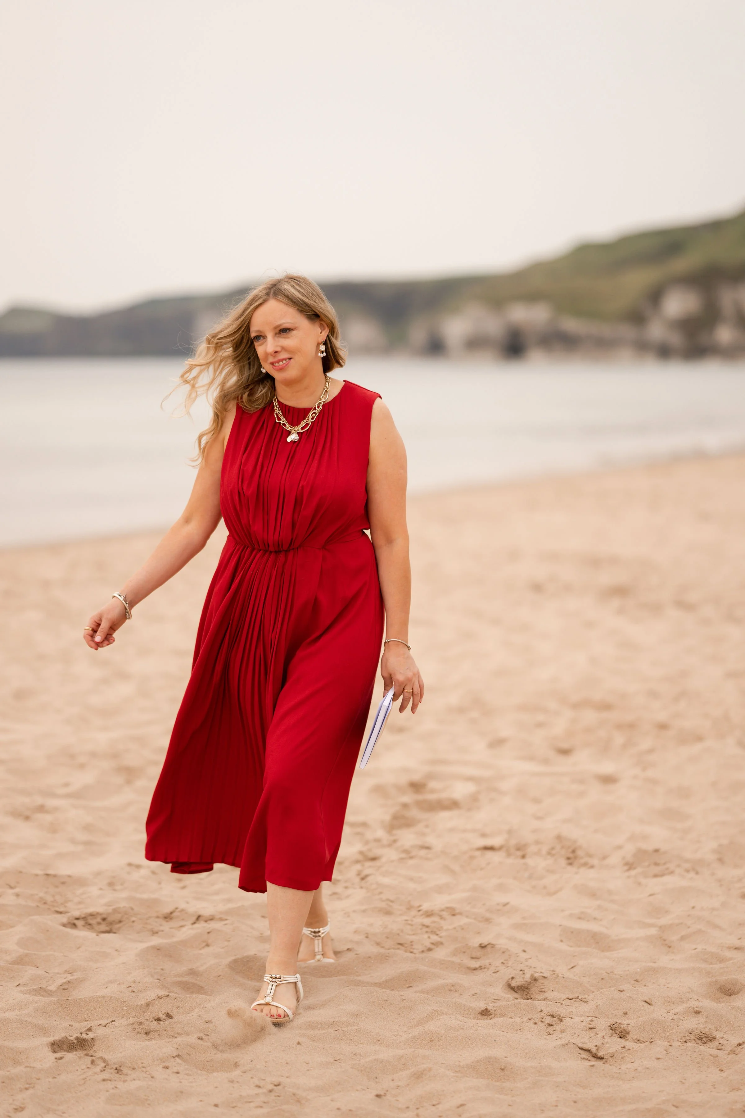 A woman in a red dress and high-heeled sandals walking on the beach with ocean and cliffs in the background.