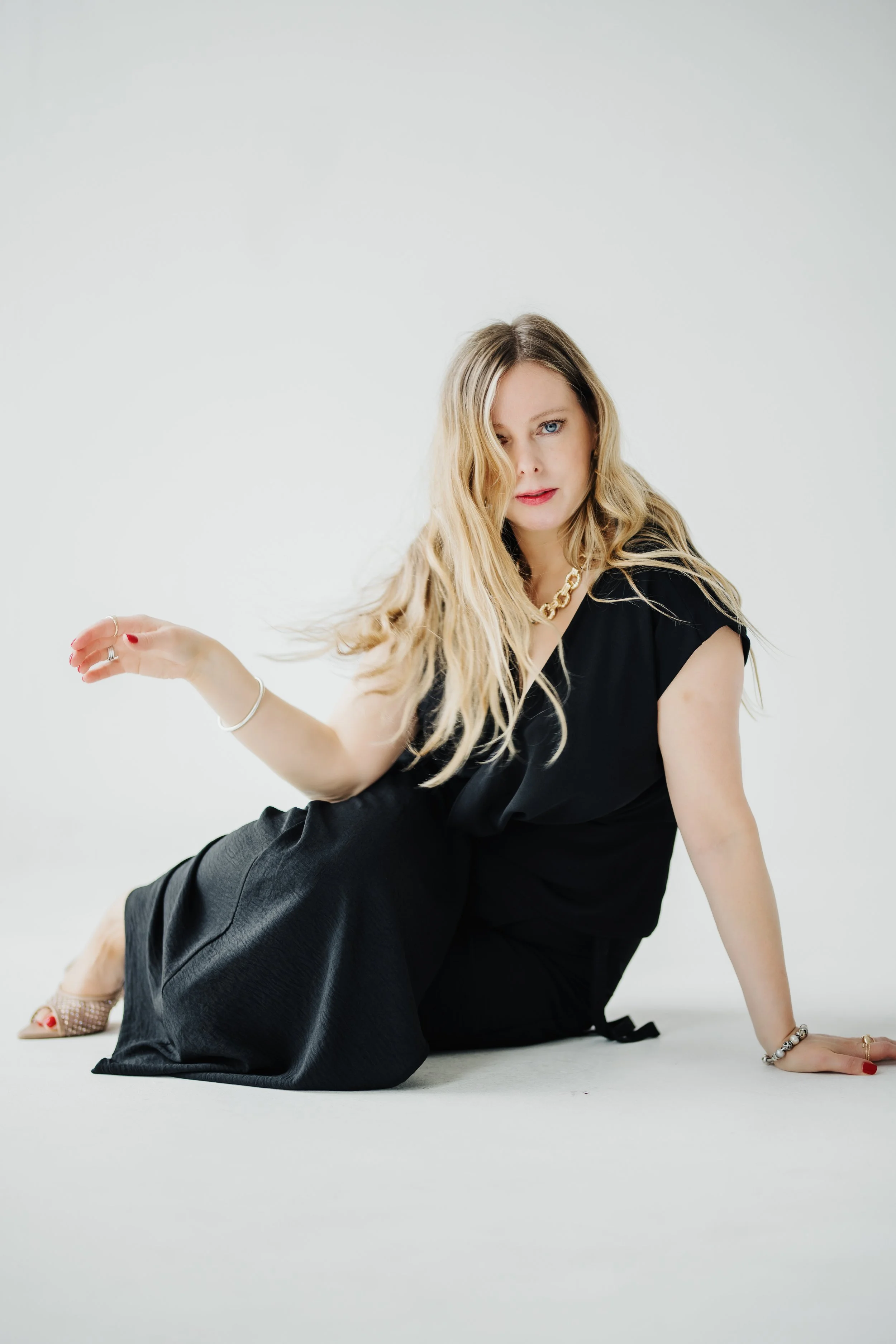A woman with blonde hair, blue eyes, wearing a black dress and gold necklace, sitting on the floor with one hand on the ground and the other raised, against a white background.