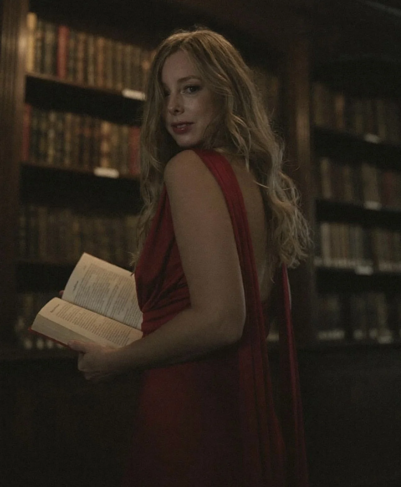 A woman with long, wavy blonde hair wearing a red dress holding an open book in a dimly lit library with dark wooden bookshelves filled with books.