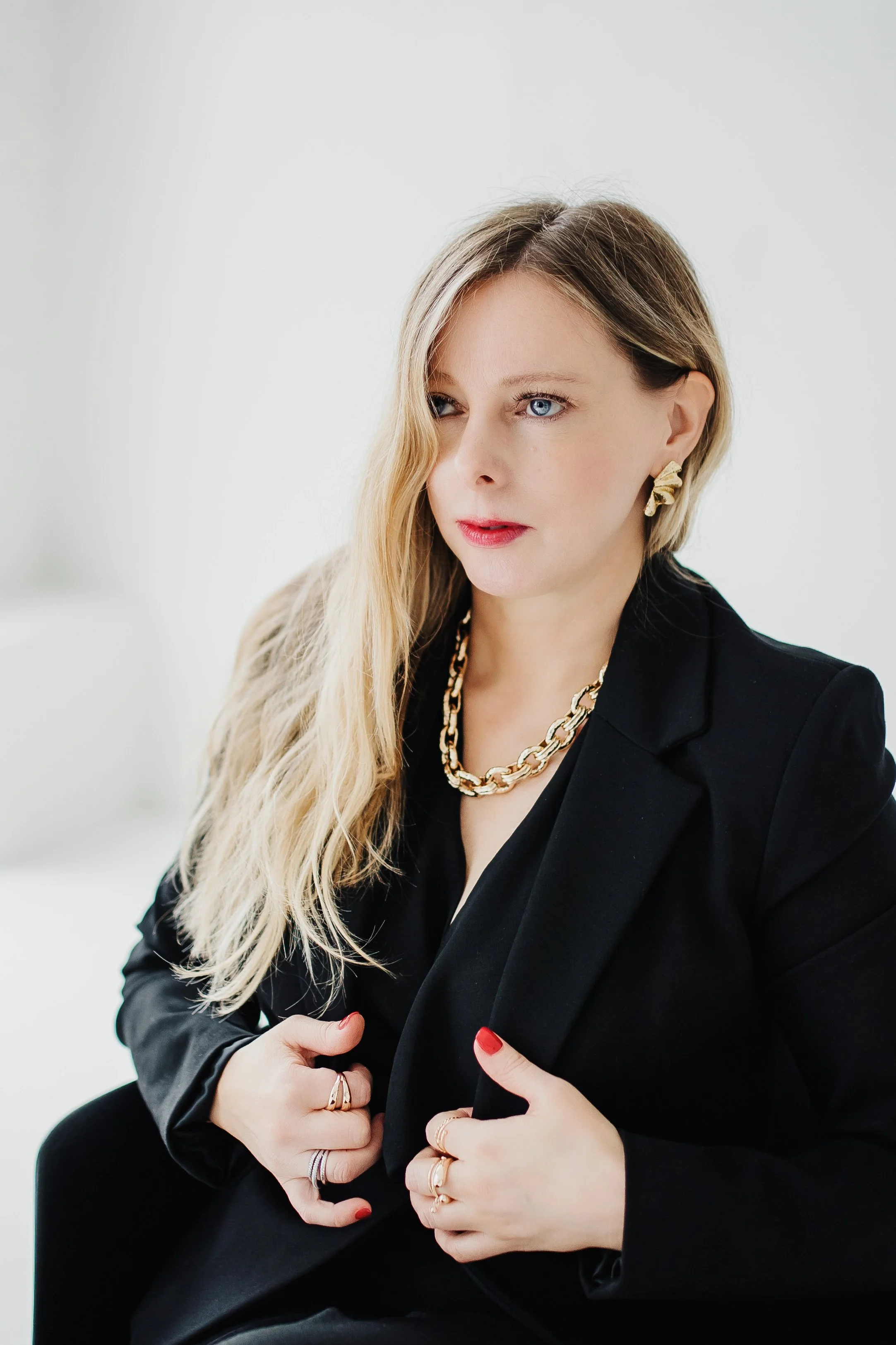 A woman with long blonde hair and blue eyes wearing a black blazer, gold jewelry including earrings, a necklace, and multiple rings, sitting against a plain light-colored background.