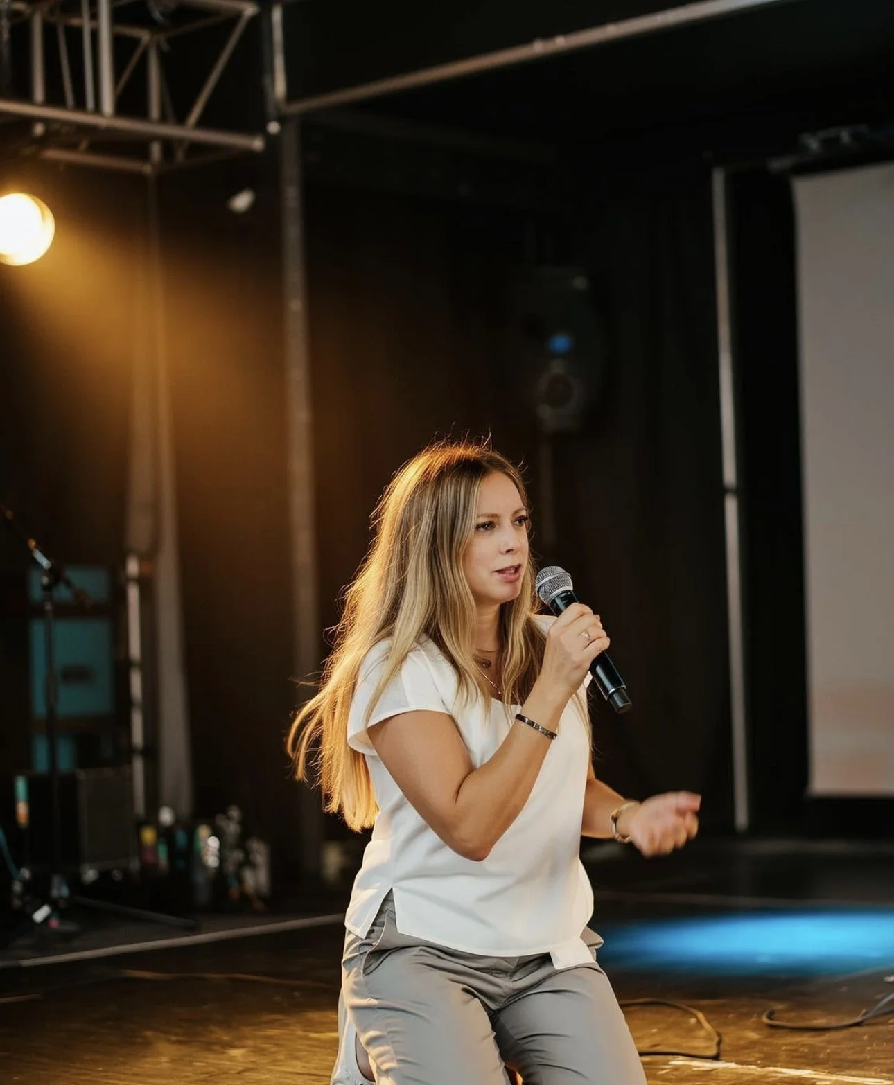 A woman with long blonde hair, wearing a white top and gray pants, speaking into a microphone during a presentation or event in a dimly lit room with stage lighting and audio equipment.