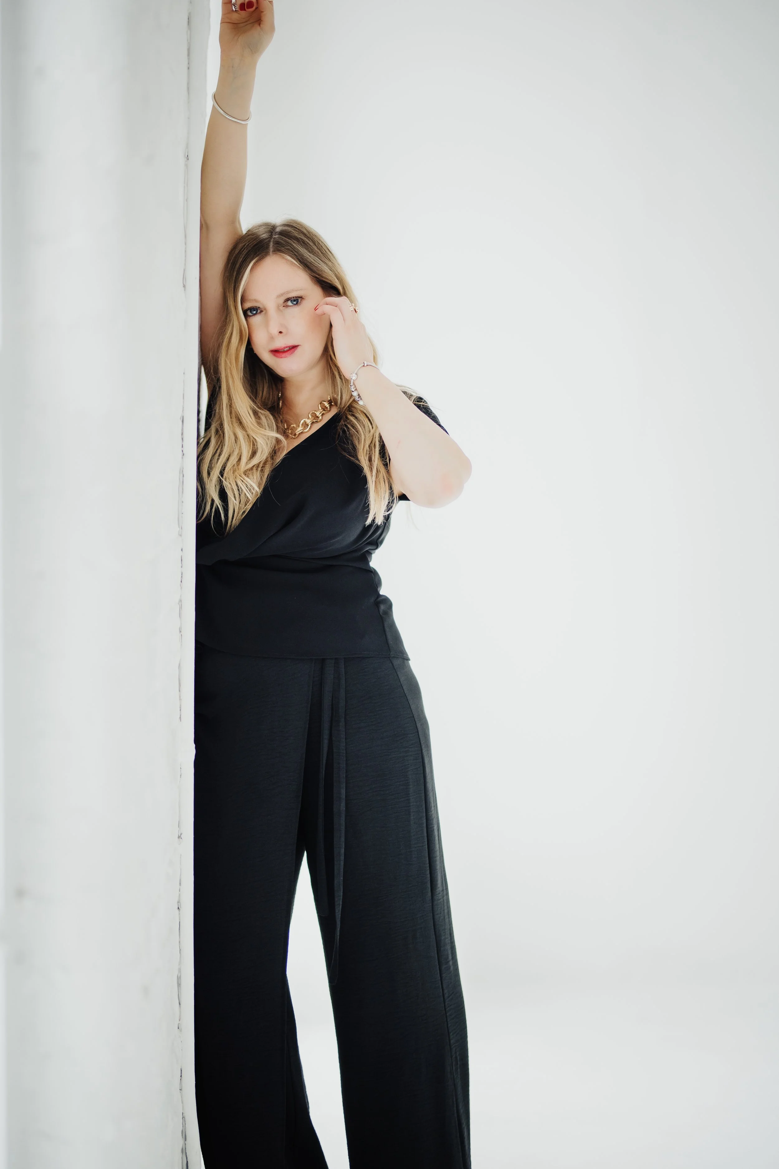 Woman in black clothing leaning against a white wall with her arm raised above her head, touching the wall, with her other hand near her face.