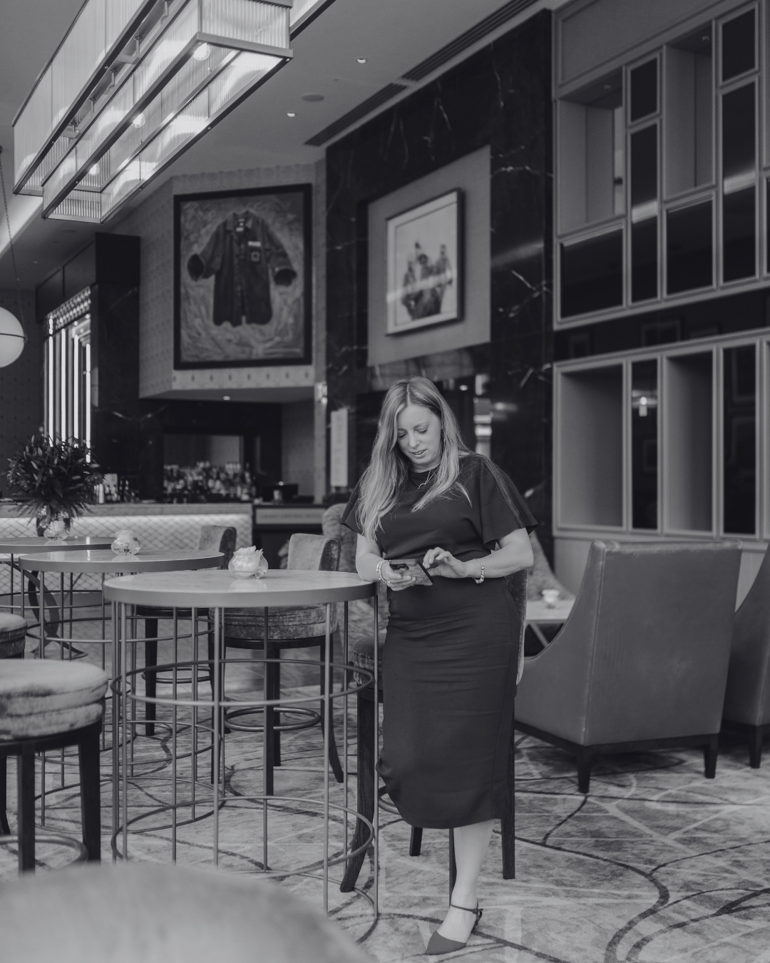 A woman in a black dress standing inside a modern, upscale lounge or restaurant, using her smartphone. The space features round tables with chairs, decorative wall art, and elegant lighting.