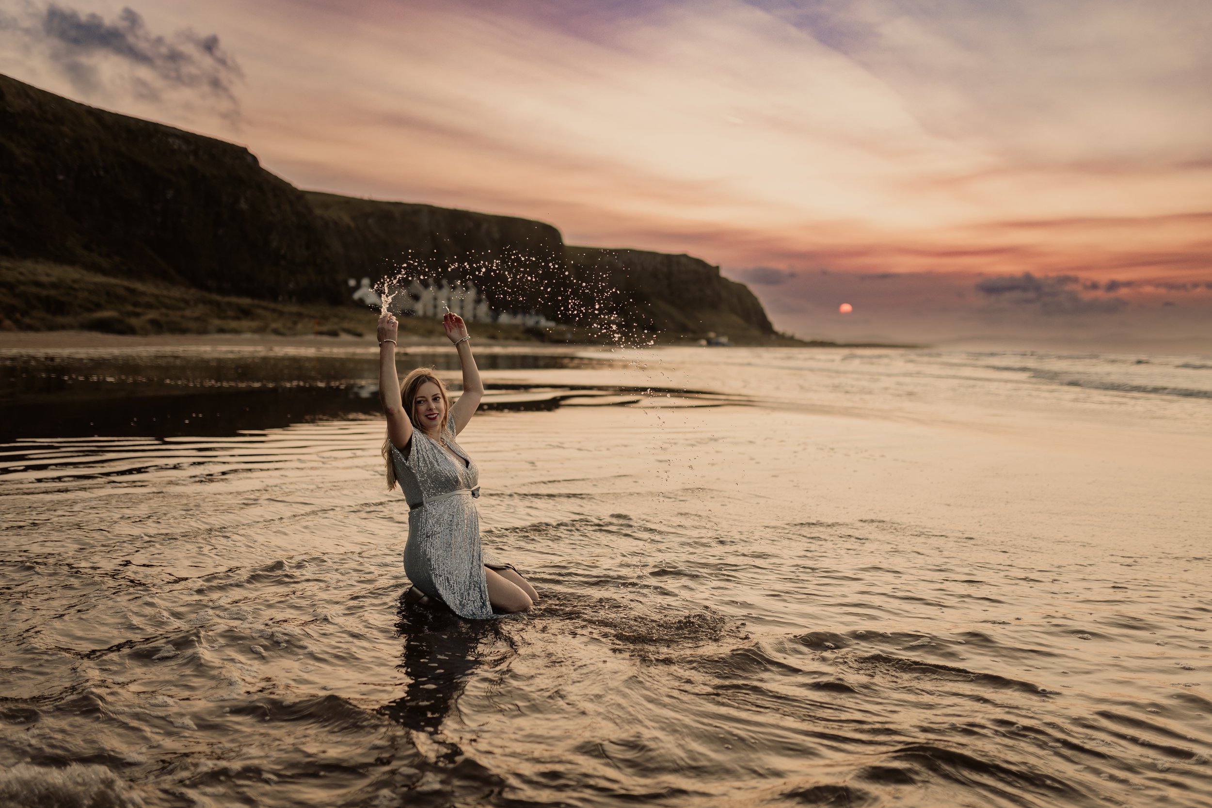 A woman kneeling in the water at sunset on a beach, raising her arms while splashing water, with cliffs in the background and a colorful sky.