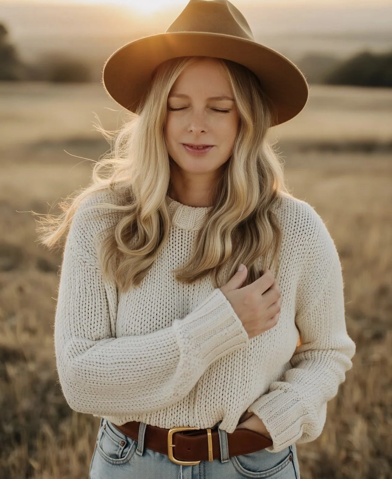 A woman with long blonde hair wearing a wide-brimmed brown hat and a cream knitted sweater, standing outdoors in a field at sunset, with her eyes closed and one hand over her heart.
