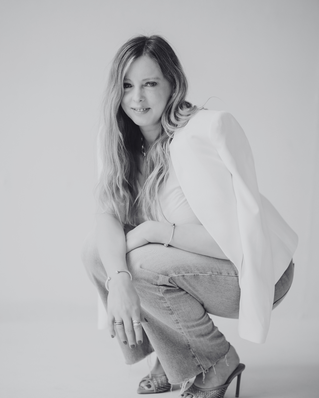 Black and white photo of a woman with long wavy hair, squatting with her arms resting on her knees, wearing a blazer, jeans, and high heels.