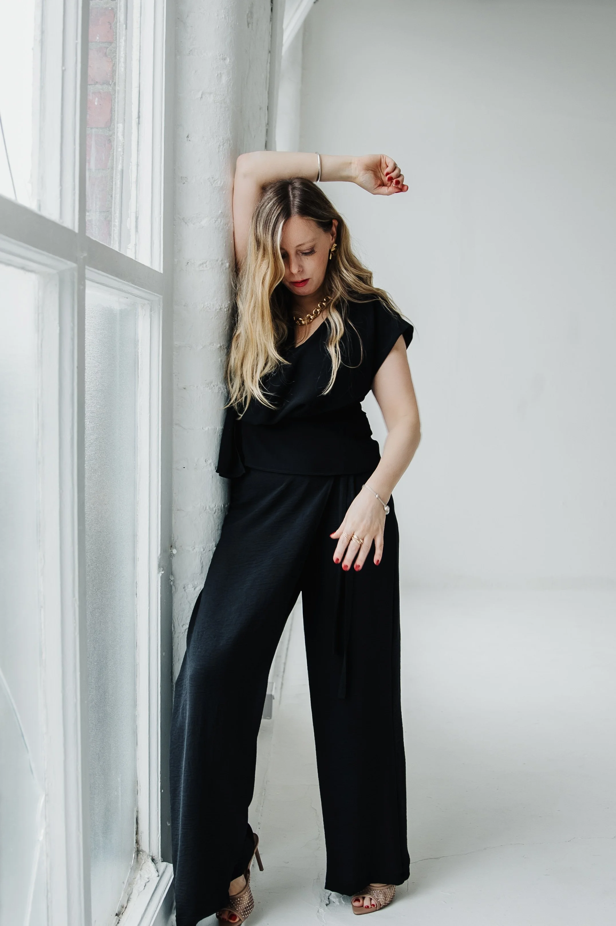 A woman in all black stylish outfit standing indoors near a white brick wall and window, with her arm raised above her head.