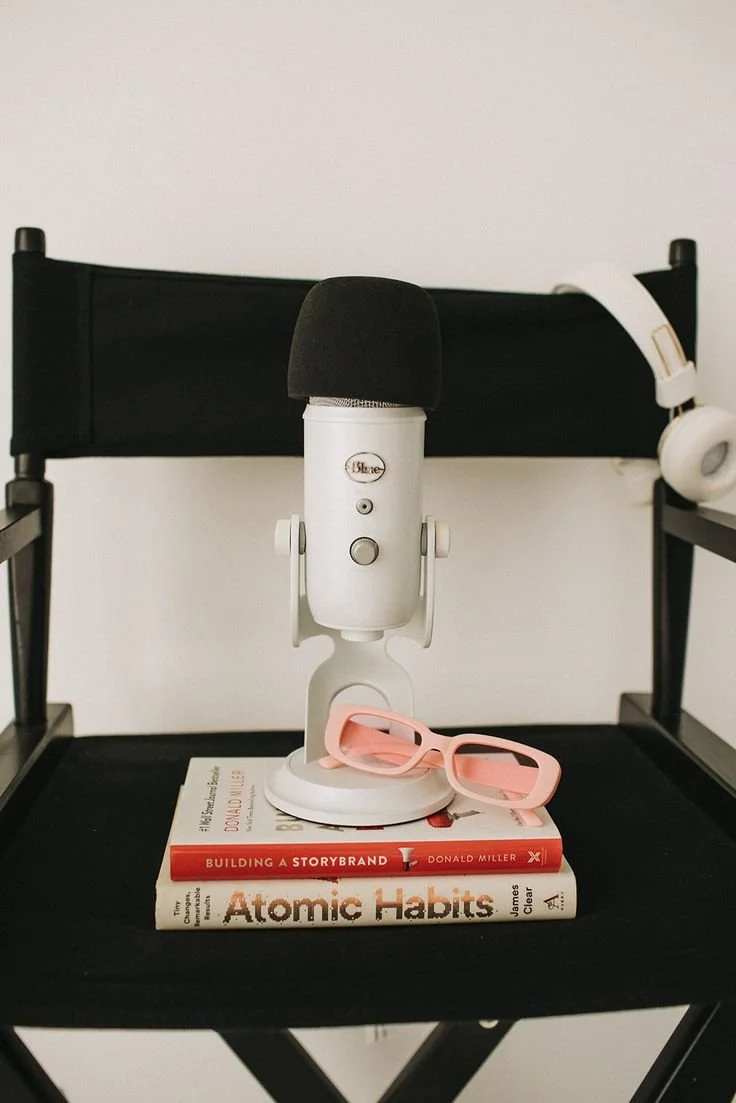 A podcasting setup with a white Blue Yeti microphone with a black windscreen, pink glasses, two books titled 'Building a StoryBrand' and 'Atomic Habits', and a pair of white headphones hanging on a black chair.