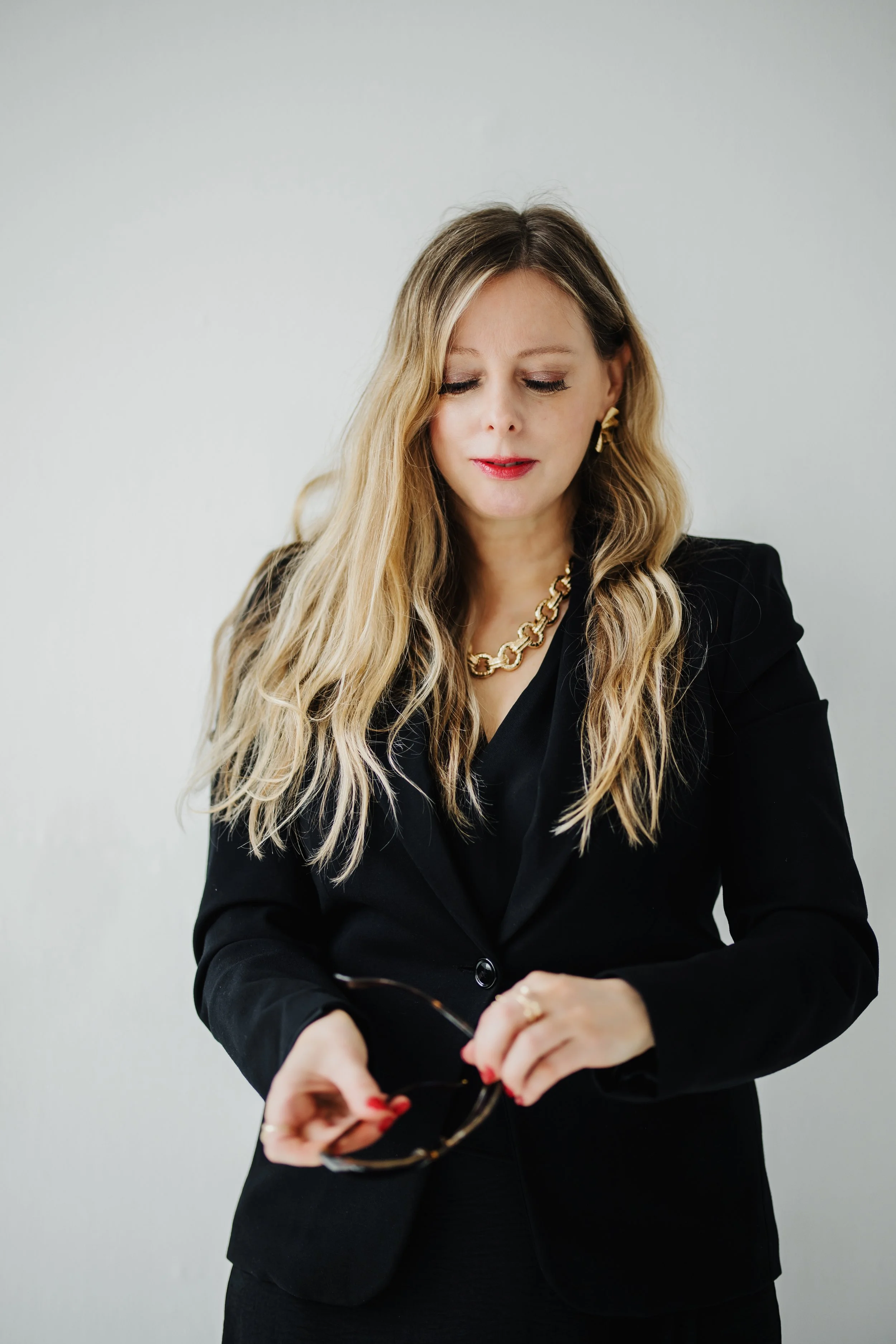 Elegant woman with long blond hair wearing a black blazer and gold jewelry, holding sunglasses and looking down.
