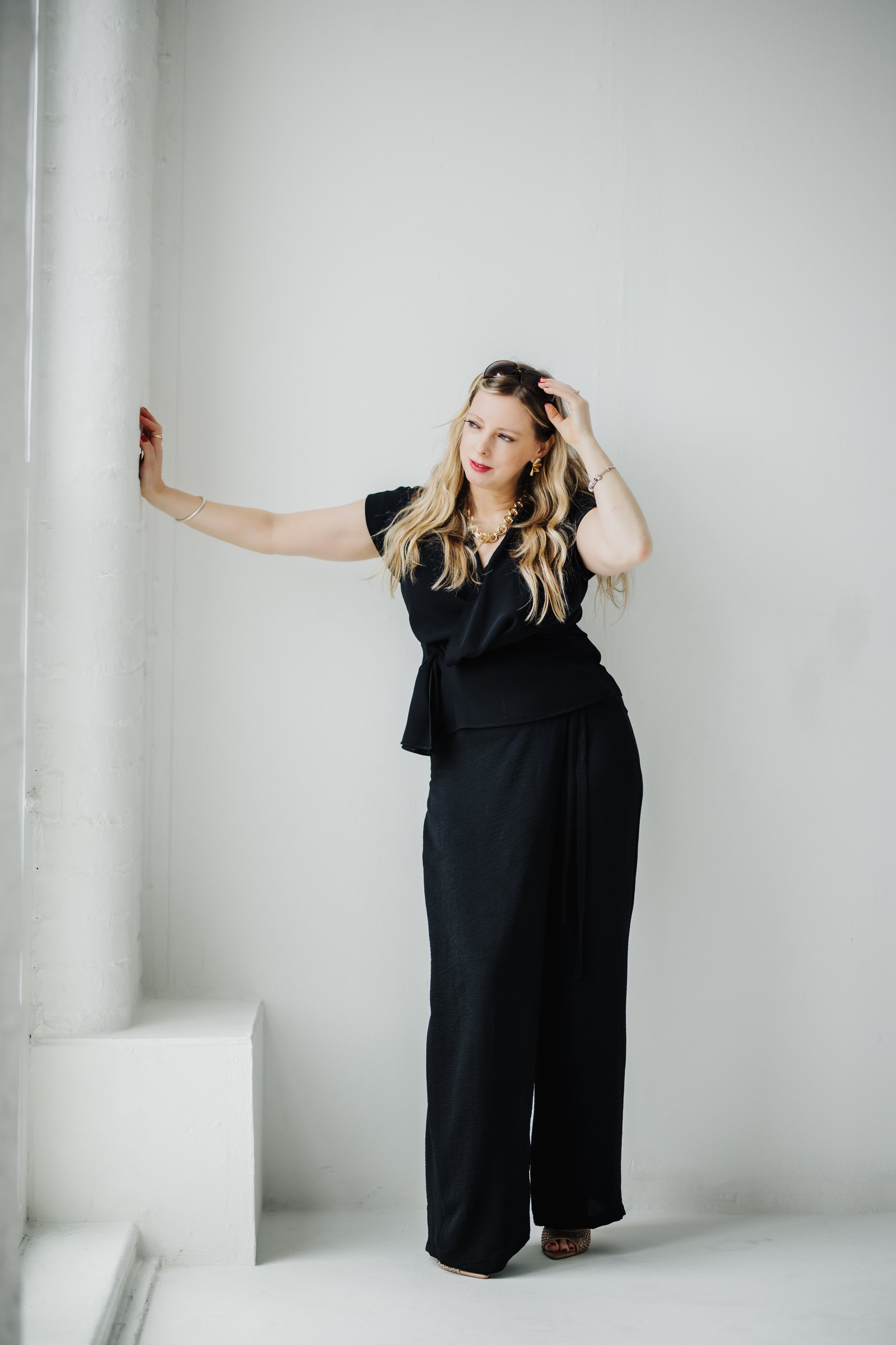A woman with long blonde hair wearing a black outfit, standing against a white wall with her right hand on a column and her left hand on her head.