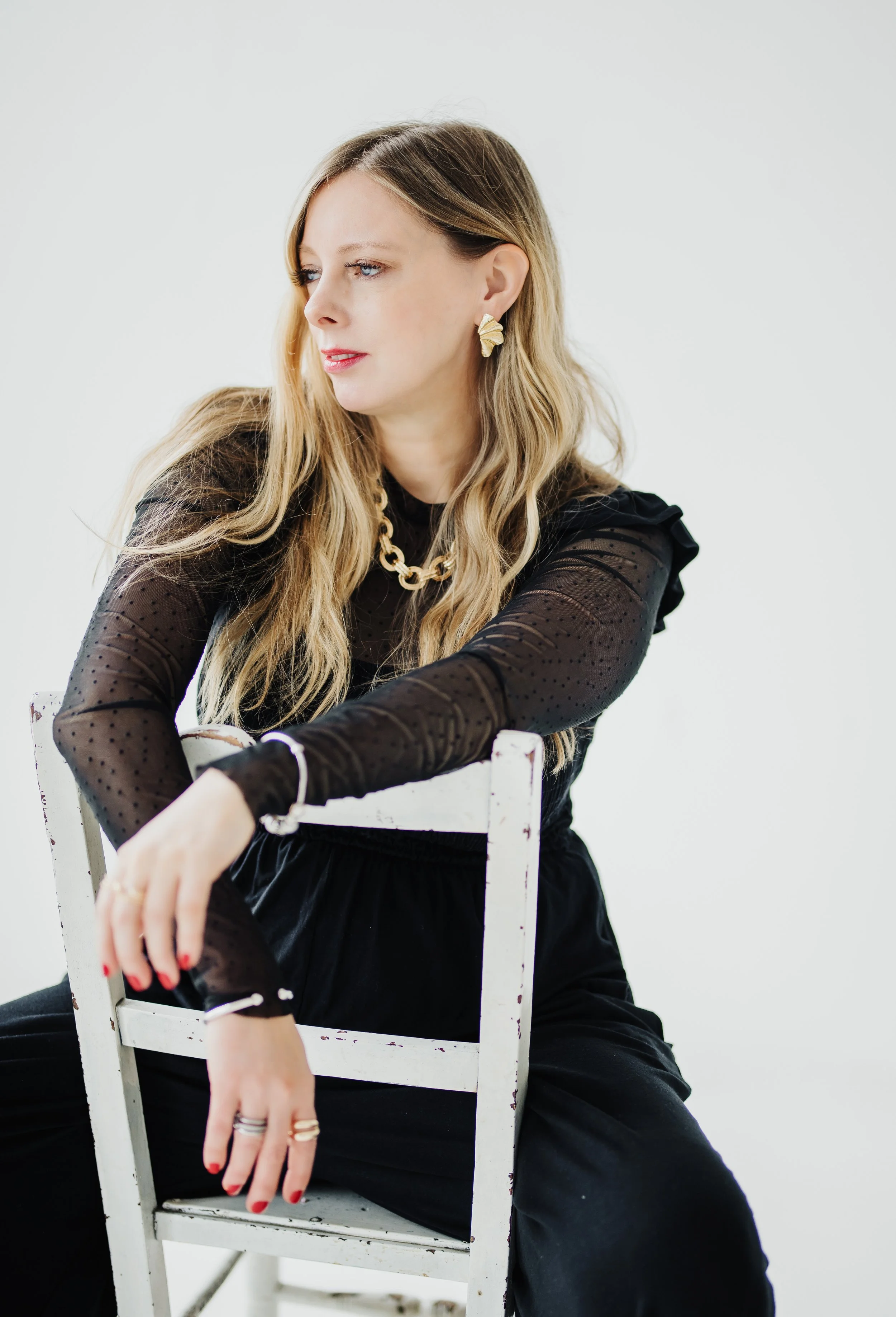 A woman with long blonde hair, wearing a black sheer polka-dot top with ruffled sleeves, black pants, gold jewelry, and red nail polish, sits on a white vintage chair against a plain white background.