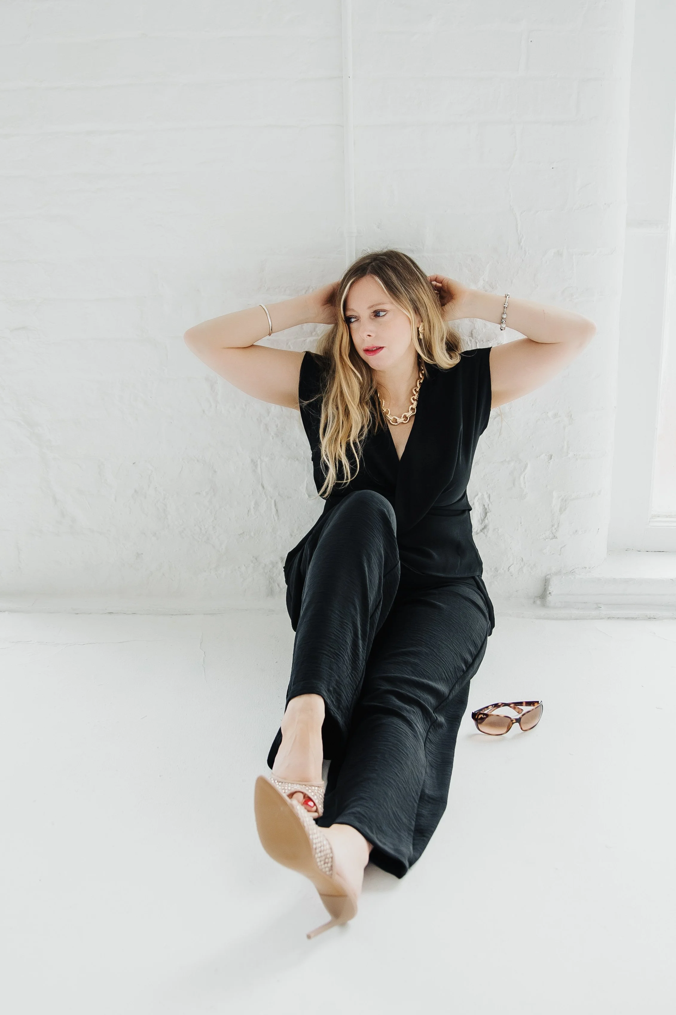 A woman in black attire sitting on the floor with one leg bent and the other stretched out, with sunglasses on the floor next to her, against a white brick wall background.