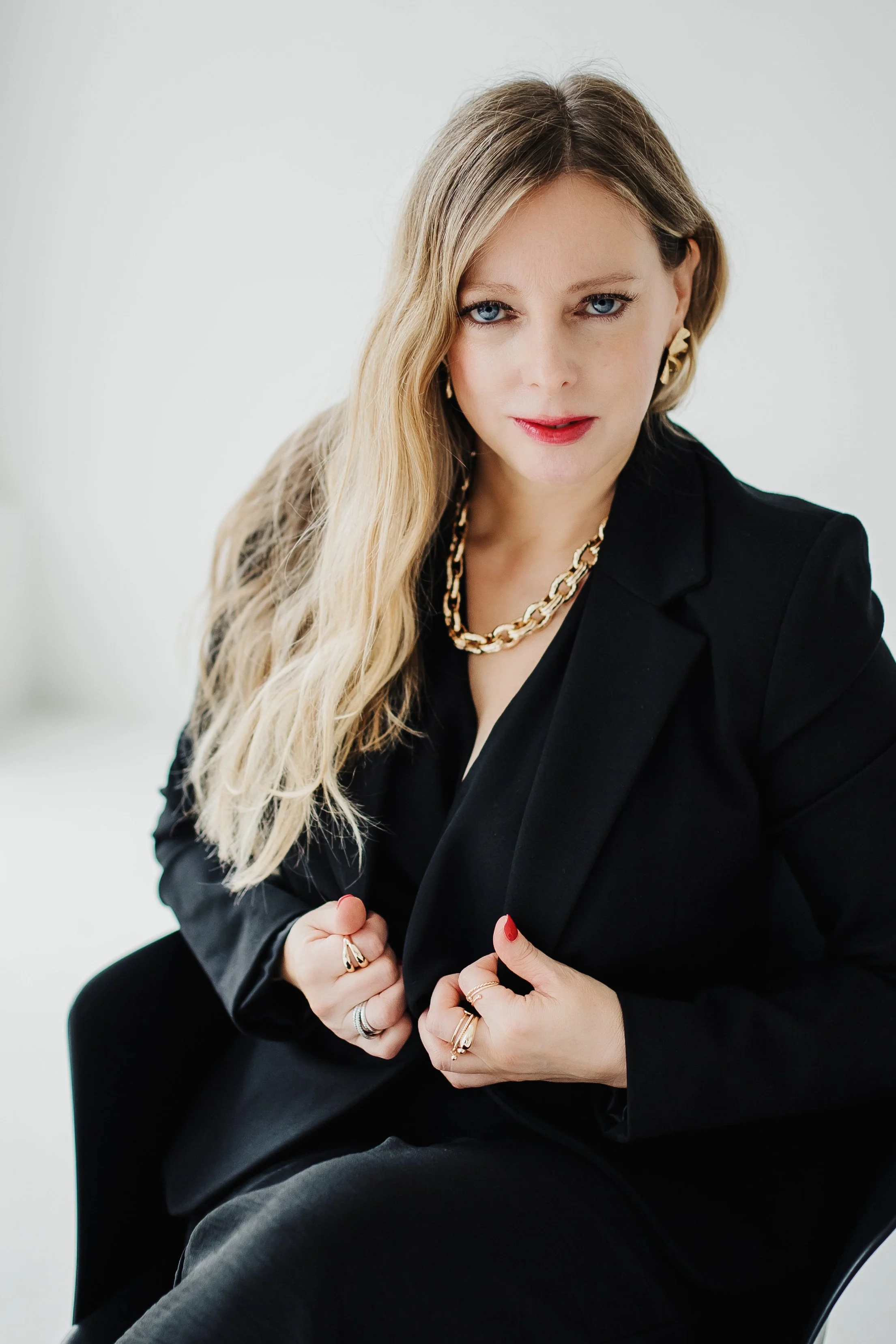 A woman with long blonde hair, blue eyes, and red lipstick, wearing a black blazer and gold jewelry, sitting and looking directly at the camera.