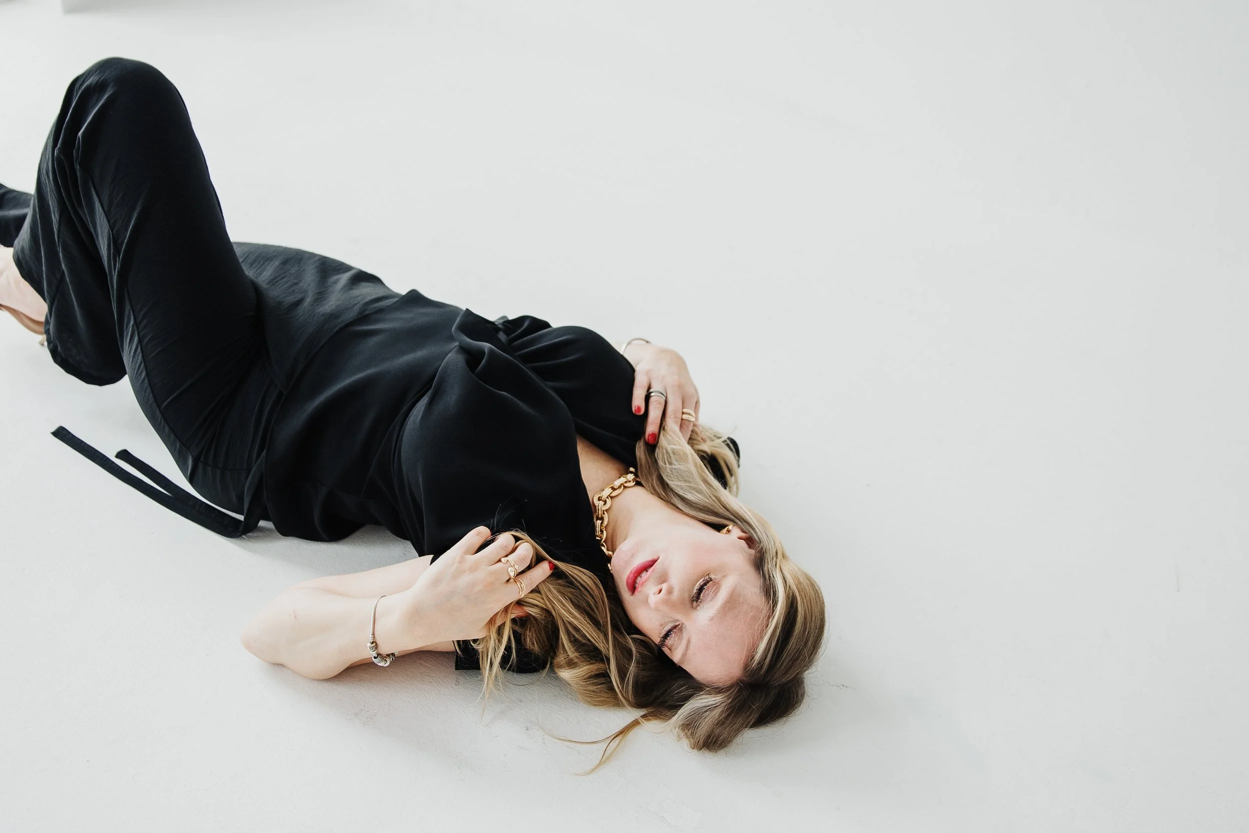 A woman lying on her side on a white surface, eyes closed, wearing a black satin dress, gold necklace, and jewelry.