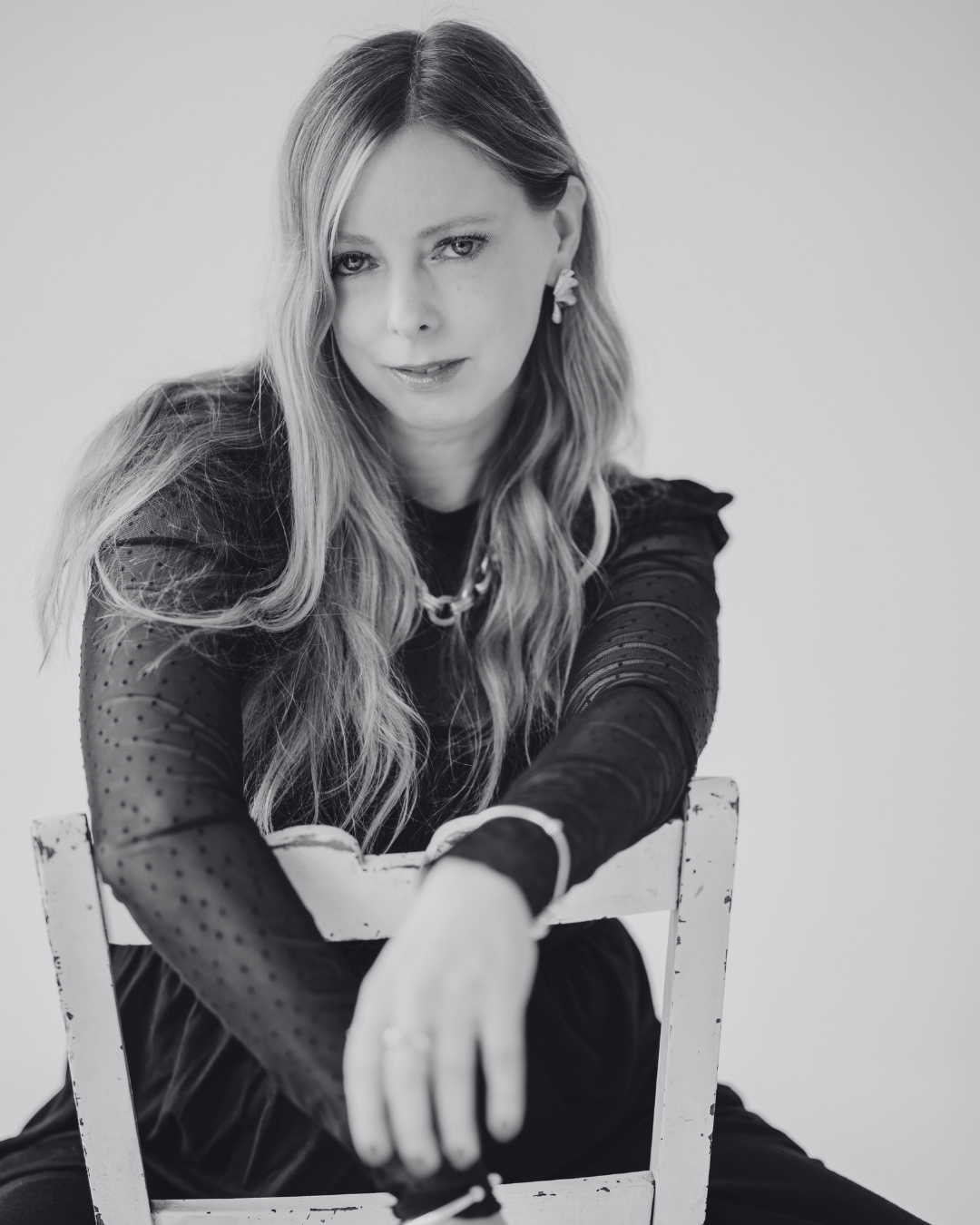 Black and white portrait of a woman with long wavy hair, wearing a dark top with sheer, dotted sleeves, sitting on a white chair and leaning forward, looking at the camera with a slight smile.