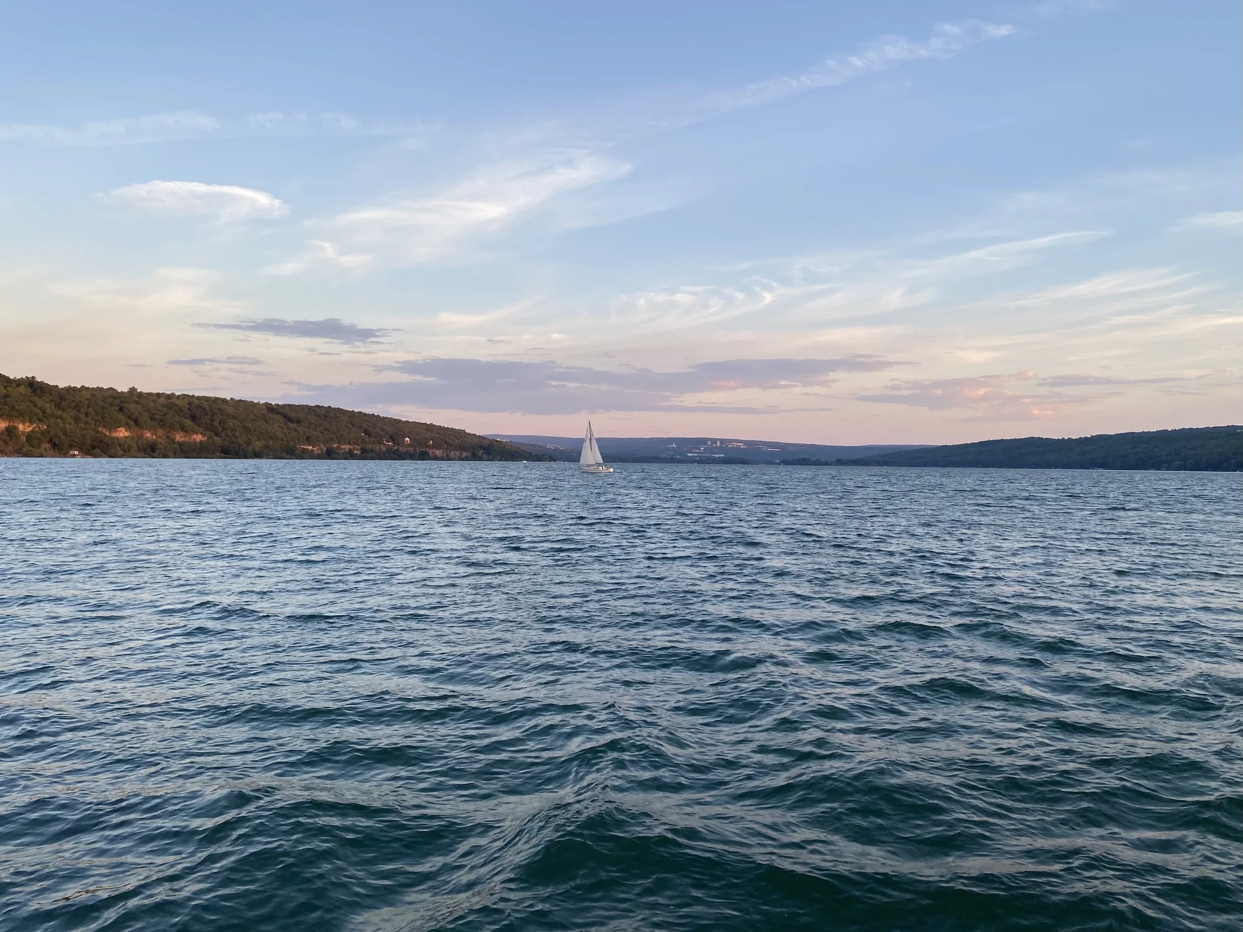 A sailboat sailing on a lake with forested hills and a partly cloudy sky in the background.