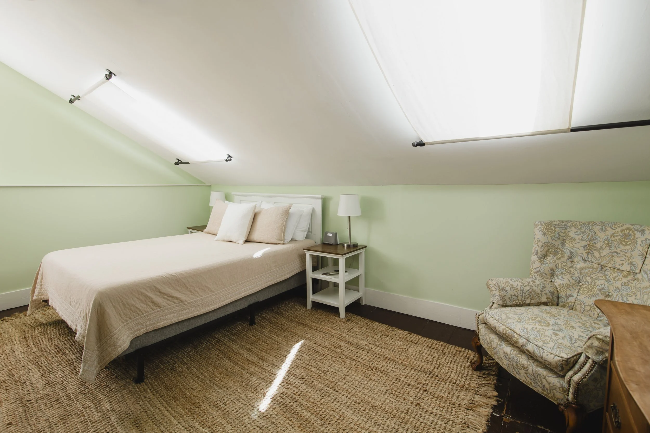 A bedroom with green walls, a white bed with beige bedding and pillows, a nightstand with a lamp and clock, an armchair with floral upholstery, a carpeted floor, and a sloped ceiling with lighting fixtures.
