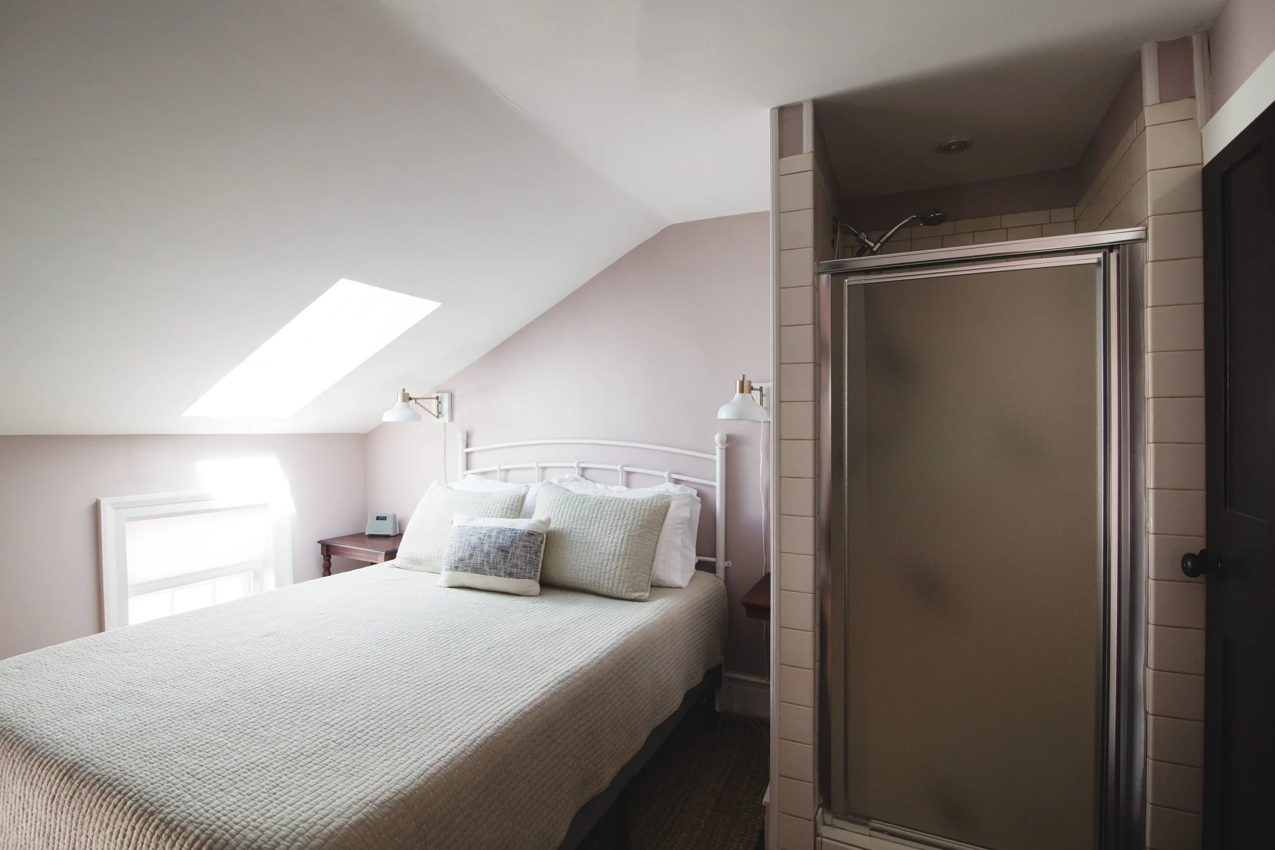 A bedroom with a sloped ceiling, skylight window, bed with pillows, and a small nightstand with a clock, adjacent to a tiled shower stall with a sliding door.