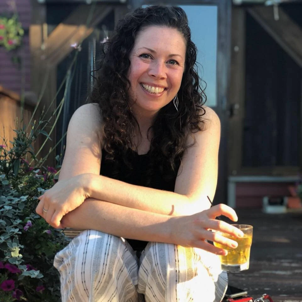 Owner and host, Jen,  with curly dark hair smiling outdoors, sitting with her arms crossed on her knees, holding a glass of drink with ice, in front of a house and plants.