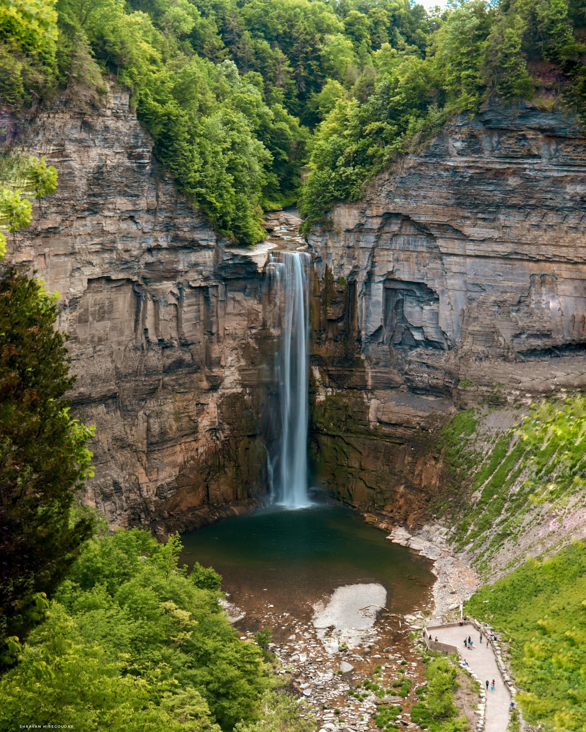 Taughannock Falls cascading down a cliff into a pool below, surrounded by lush green trees and a viewing platform with people.