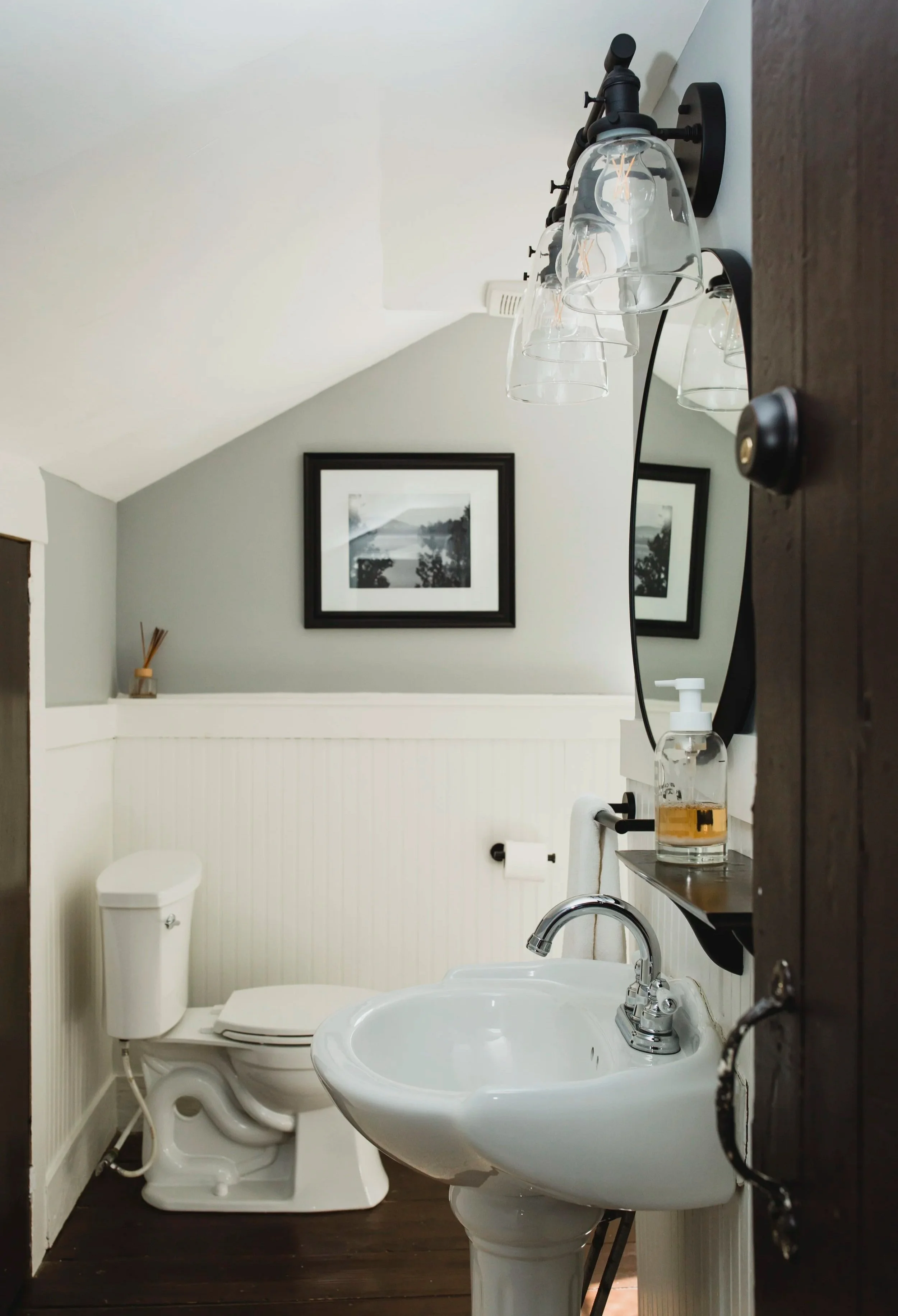 A small bathroom with a white toilet, a white sink, and a mirror. There is a black wall-mounted fixture with empty glasses and a soap dispenser on the sink. A framed black-and-white landscape photograph hangs on the wall, and a small reed diffuser is