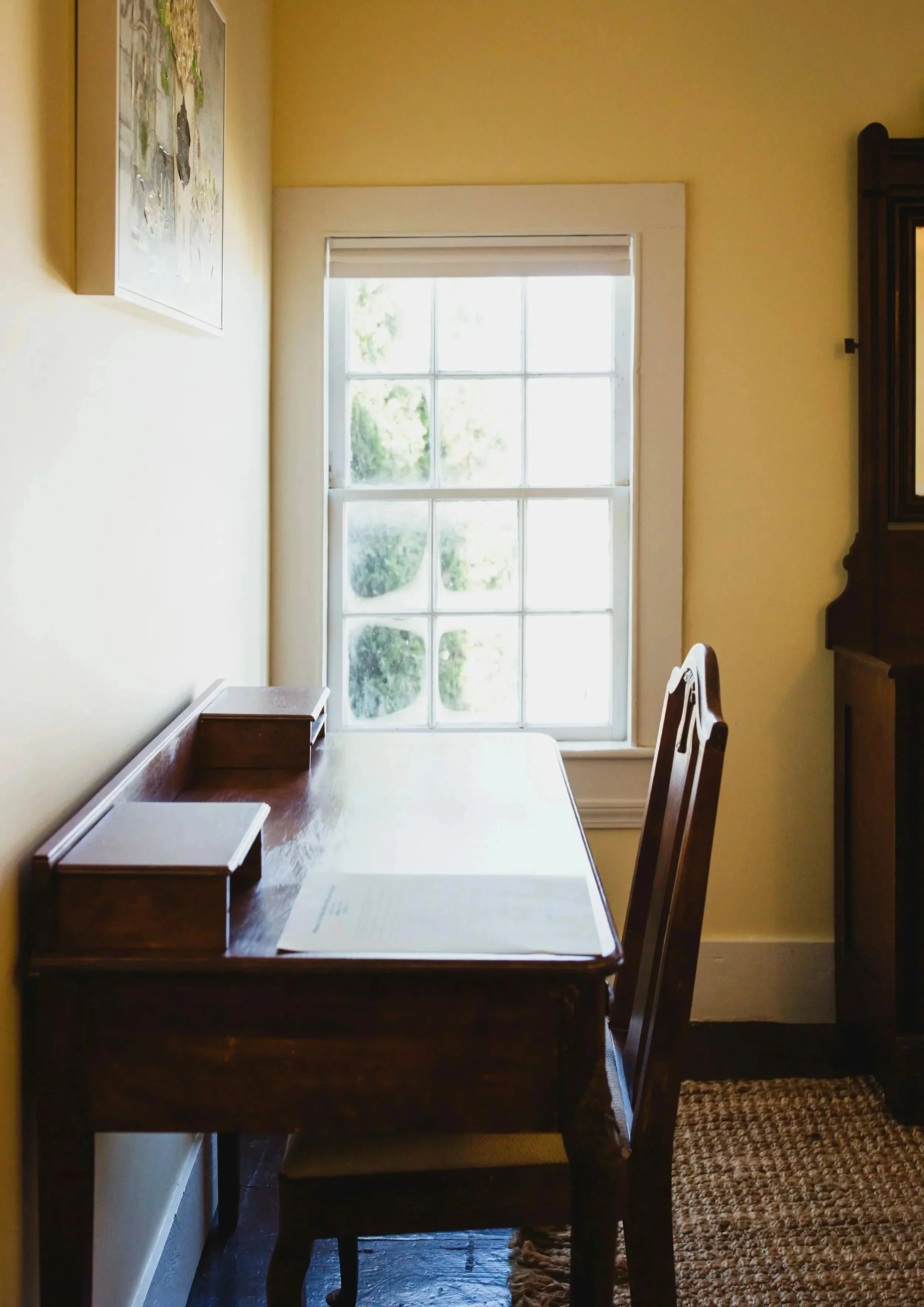 A wooden dining table with a paper and a wooden tray placed on it. There is a wooden chair to the right of the table, and a window in the background allowing sunlight into the room.
