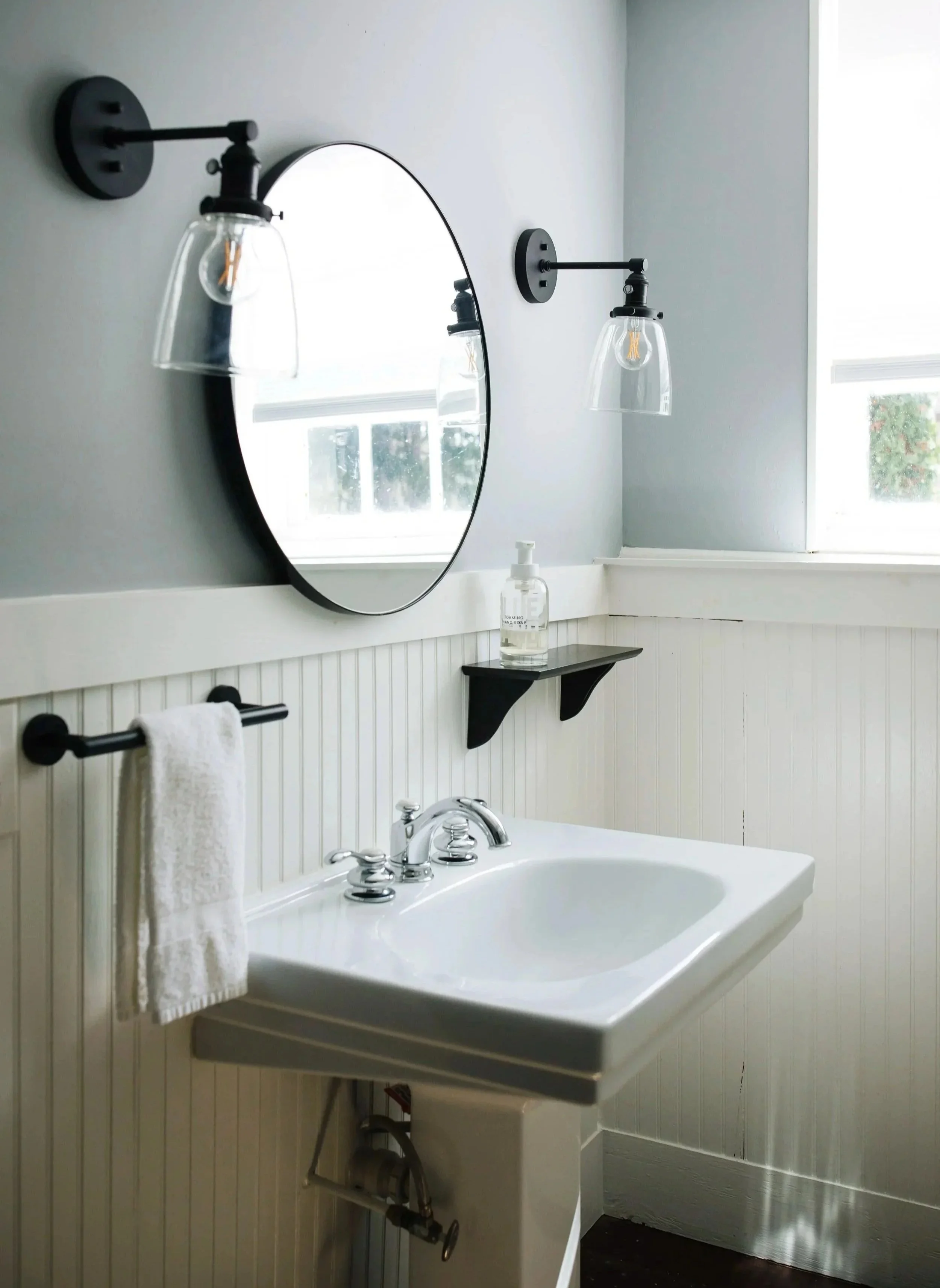 A white bathroom with a porcelain sink, a mirror above it, and two wall-mounted light fixtures on either side of the mirror. The sink has a chrome faucet and a hand soap dispenser on the side. There is a white towel hanging on a black bar below the m