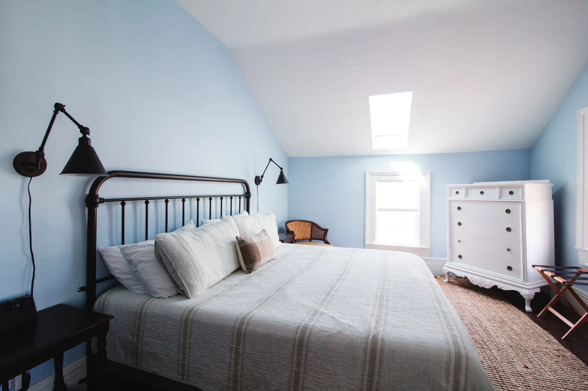 A bedroom with light blue walls, a bed with a white quilt and pillows, black wall-mounted lamps on each side, a window with white frame, a white dresser, and a brown hexagonal chair.