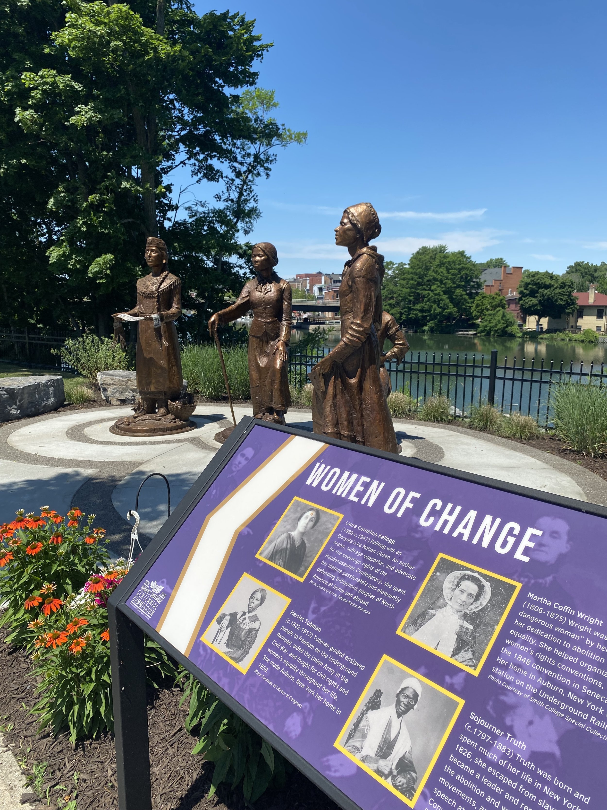 Four life-sized bronze statues depicting historic women, standing on a circular stone platform outdoors near a body of water in Seneca Falls, with a sign in the foreground titled 'Women of Change' and informational text.