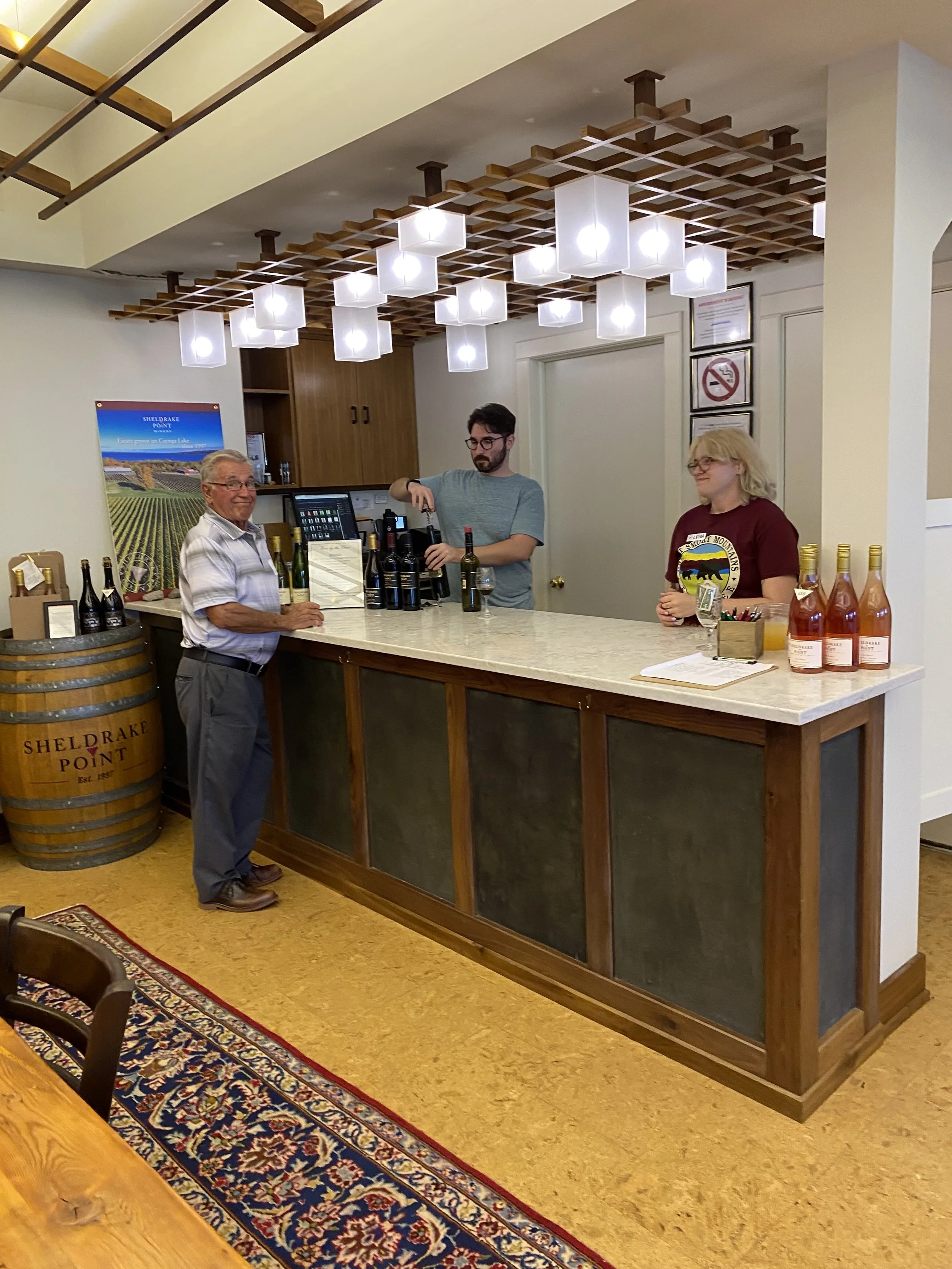 A sommelier behind a counter with two bottles of wine, two women at the counter, with wine bottles on the shelf and a poster of Sheldrake Point Vineyard in the background, inside a the winery's tasting room.