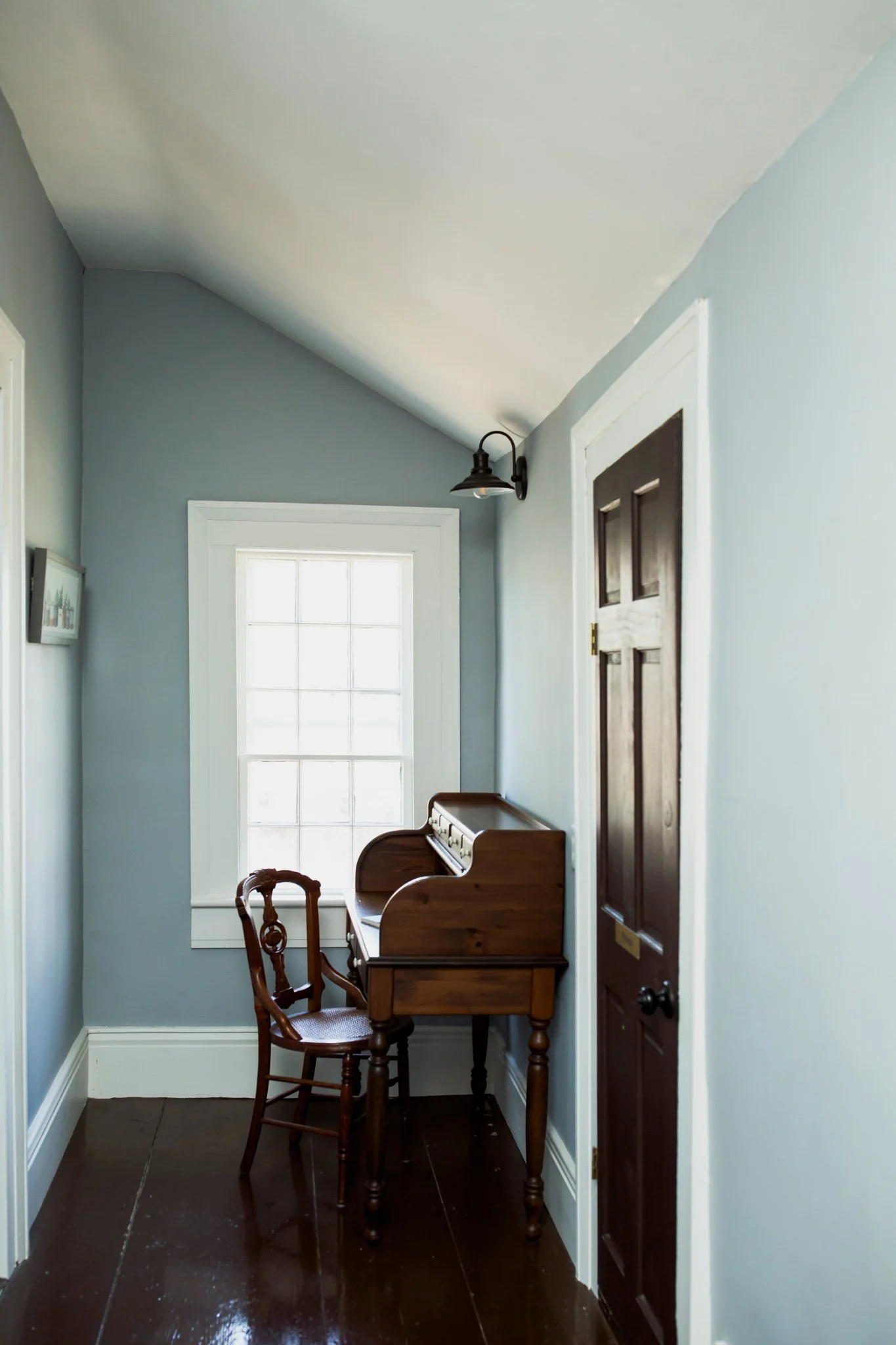 A narrow hallway with light blue walls, a white ceiling, and a dark brown wooden door on the right. There is a large window with multiple panes letting in natural light and a small, vintage wooden writing desk with a matching chair against the wall b