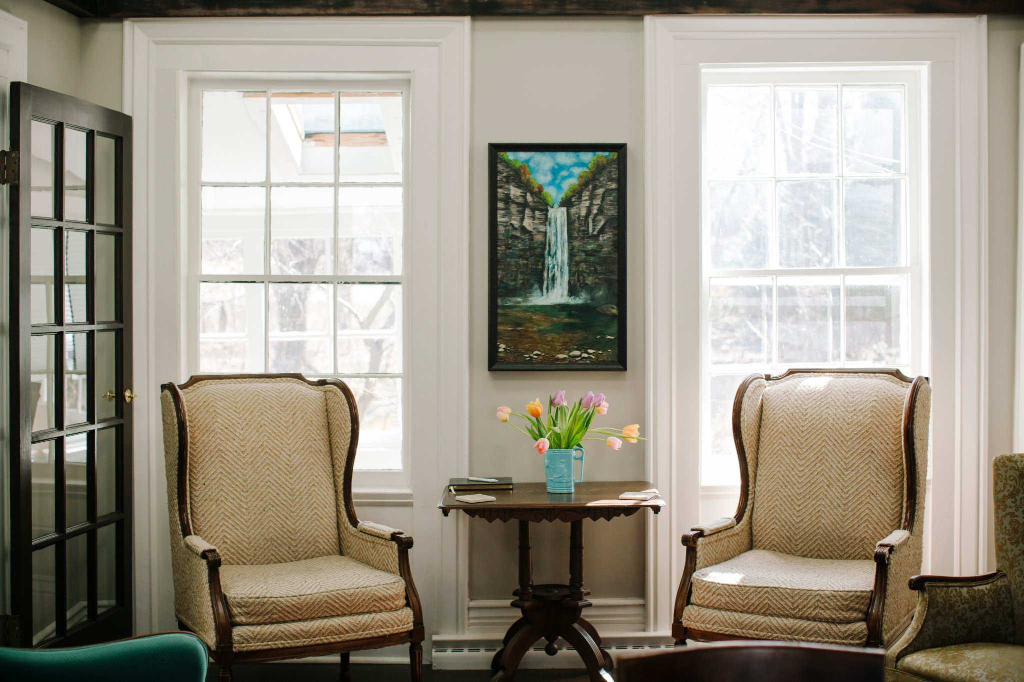 A cozy living room corner with two beige upholstered armchairs with dark wooden frames on either side of a small wooden table. The table has a blue vase with pink tulips and some books on it. Behind, large windows let in natural light, and a colorful waterfall painting hangs between the windows.