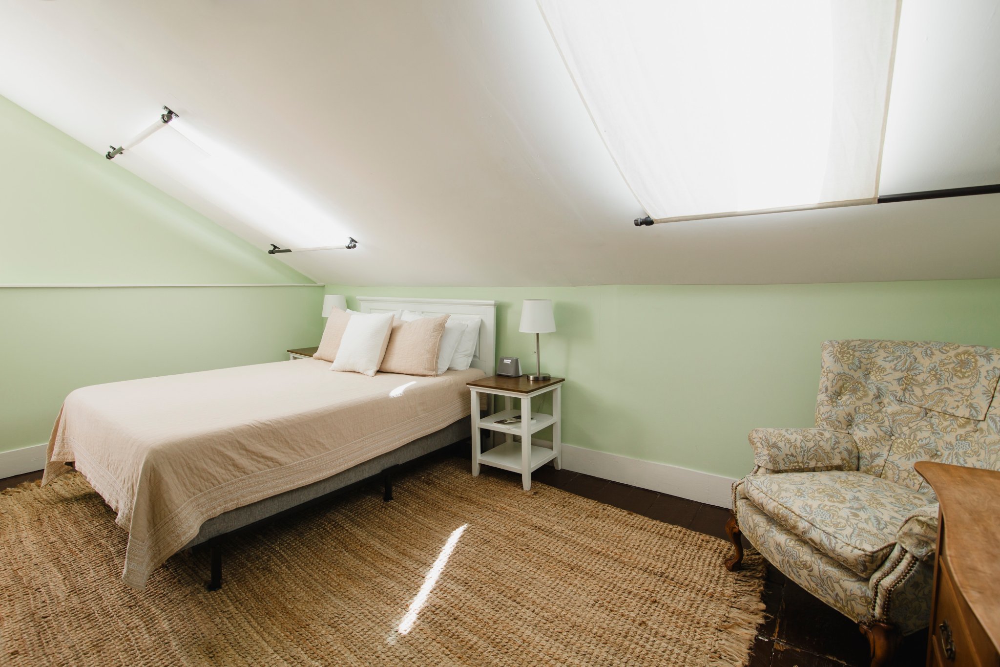A bedroom with light green walls, a bed with beige bedding and pillows, a white nightstand with a lamp and a phone, a patterned armchair, and ceiling lights.