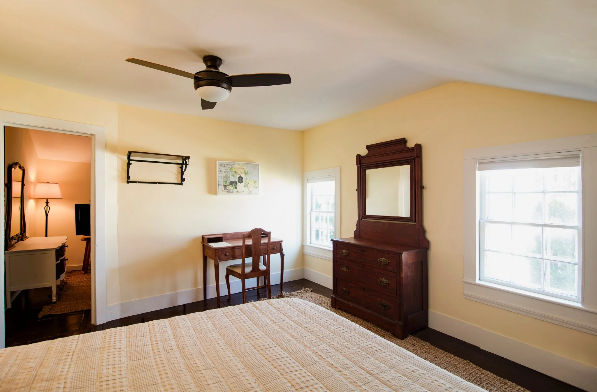 A bedroom with yellow walls, a wooden dresser with a mirror, windows letting in natural light, and a ceiling fan.
