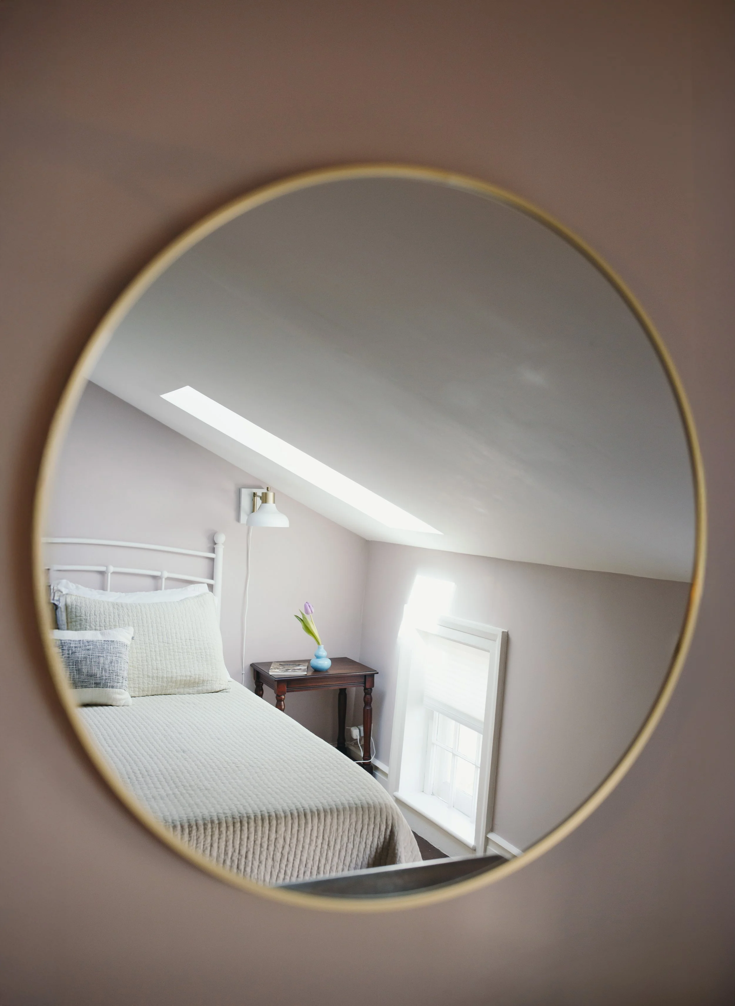 Reflection in a round mirror showing a bedroom with a bed, a bedside table with a small blue vase and a single purple tulip, wall-mounted lamp, and a window with white blinds.