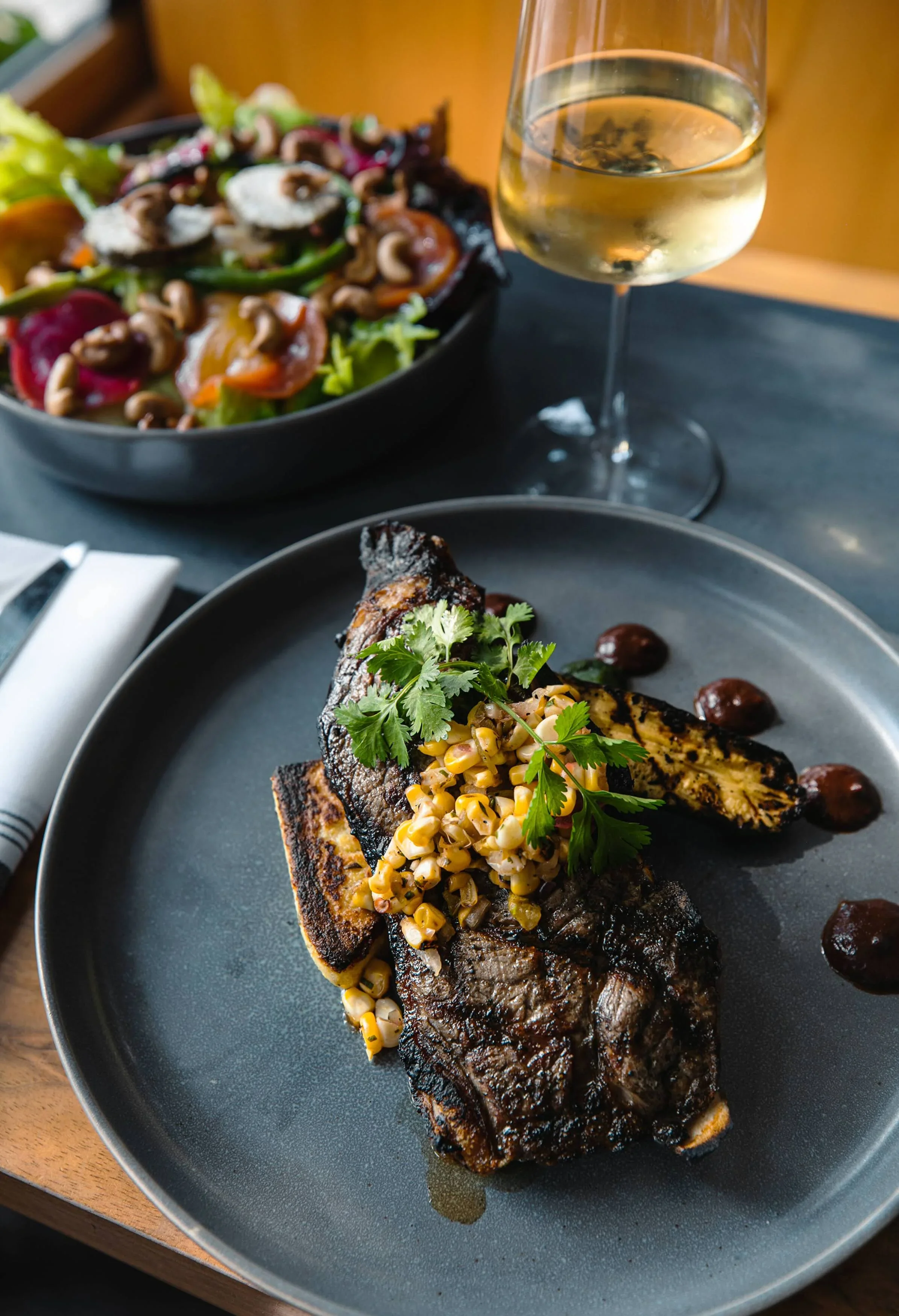 A grilled steak topped with corn salad on a black plate, garnished with cilantro, and a glass of white wine in the background.