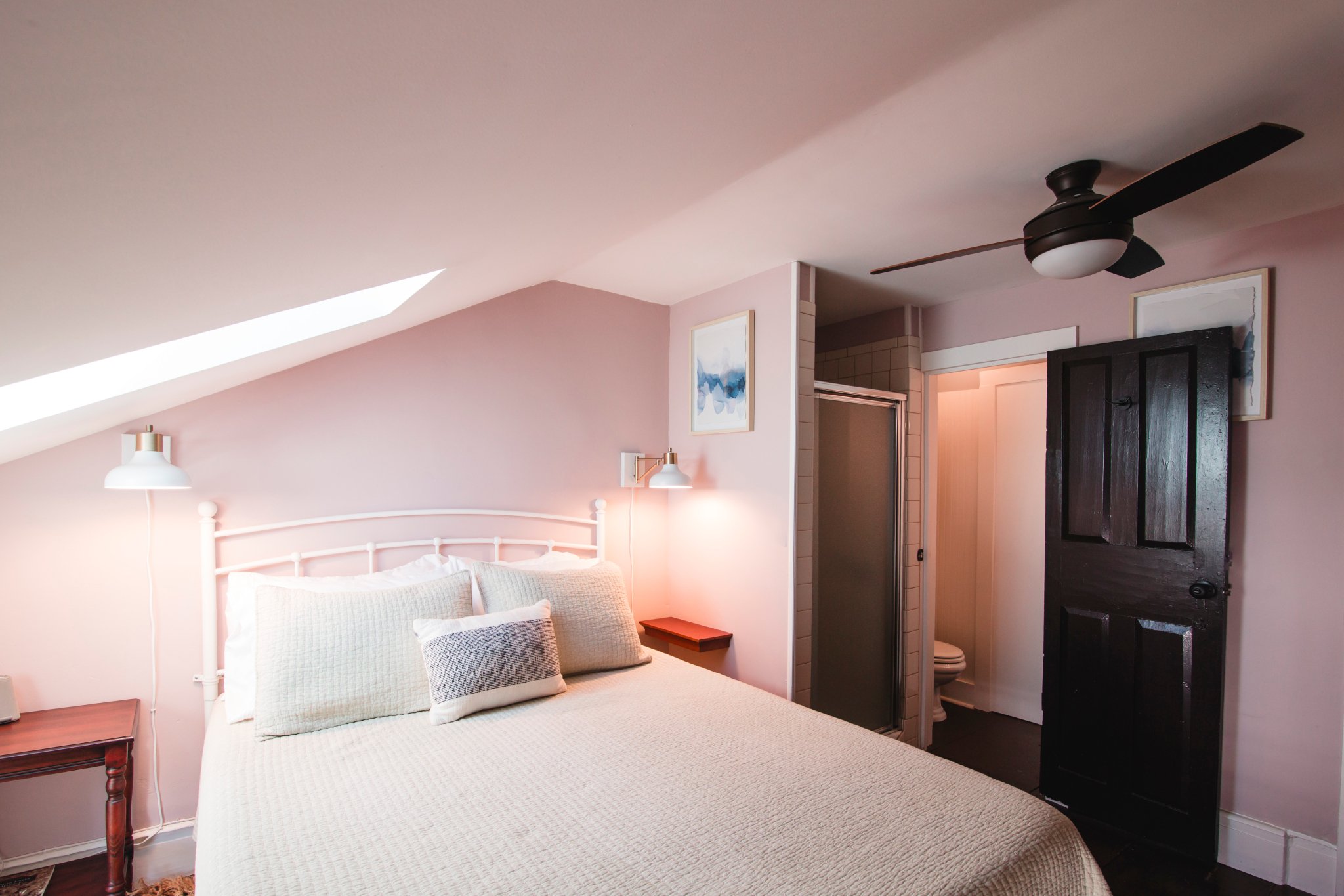 A cozy bedroom with a pink accent wall, a white bed with textured pillows, wall-mounted reading lamps, abstract art, a ceiling fan, and a doorway leading to a bathroom.
