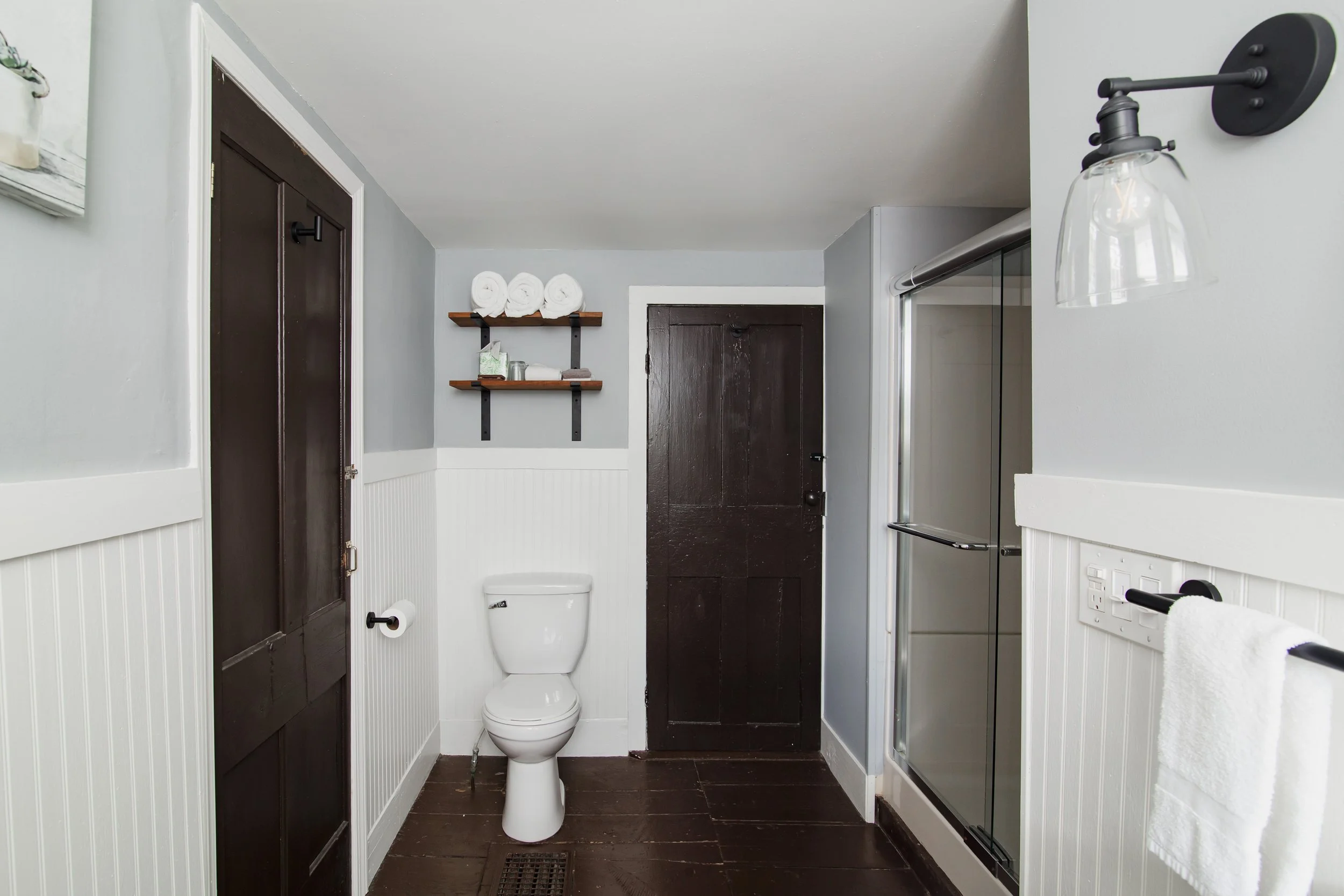 A small bathroom with dark brown door, white beadboard wainscoting, and gray walls. There is a white toilet against the back wall, with a brown wooden shelf above it holding three rolled white towels and some toiletries. To the right, there is a show