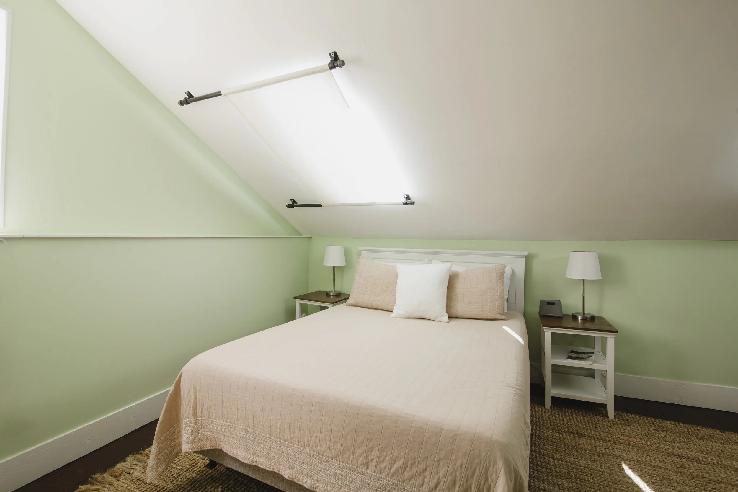 A bedroom with light green walls, a bed with beige bedding and pillows, two bedside tables with lamps, a white headboard, and a sloped ceiling with a mounted track lighting system.