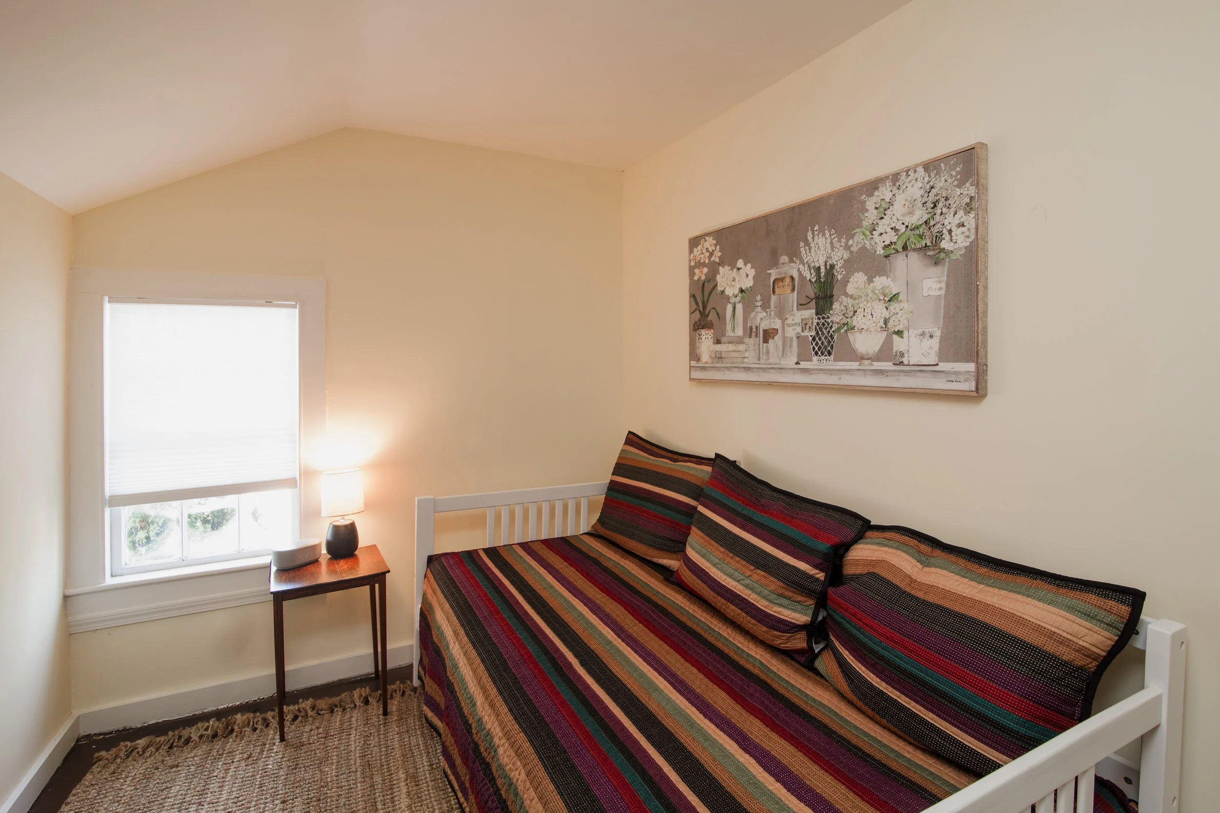 A bedroom corner with a bed that has a colorful striped bedspread and pillows, a window with blinds, a small wooden side table with a lamp, a decorative vase, and a painting of flowers on the wall.