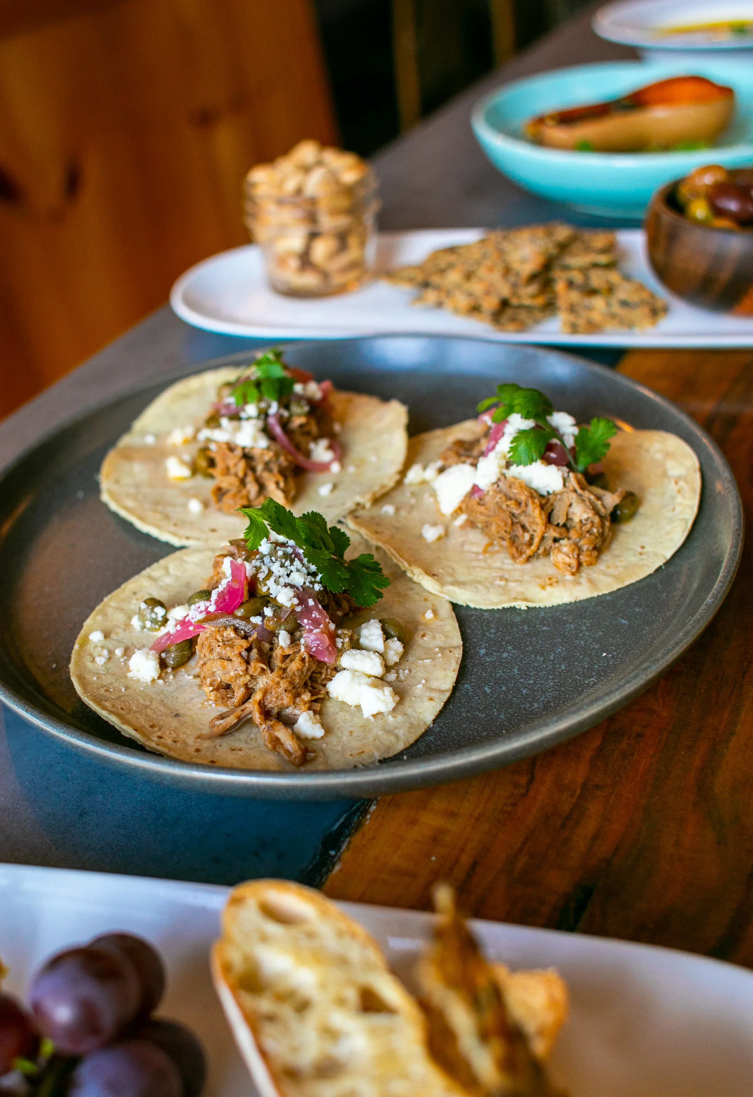 Close-up of three street-style tacos with shredded meat, cheese, onions, and cilantro on a black plate in the foreground. In the background, there are bowls of beans and other side dishes on a wooden table.