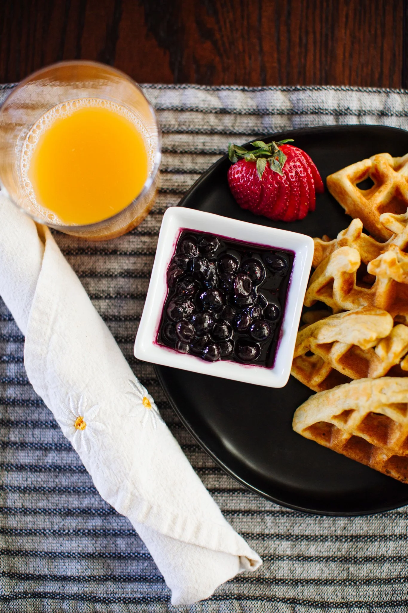 Breakfast plate with sliced strawberries, waffles, and blueberries in a bowl, with a glass of orange juice and a rolled napkin on a striped tablecloth.