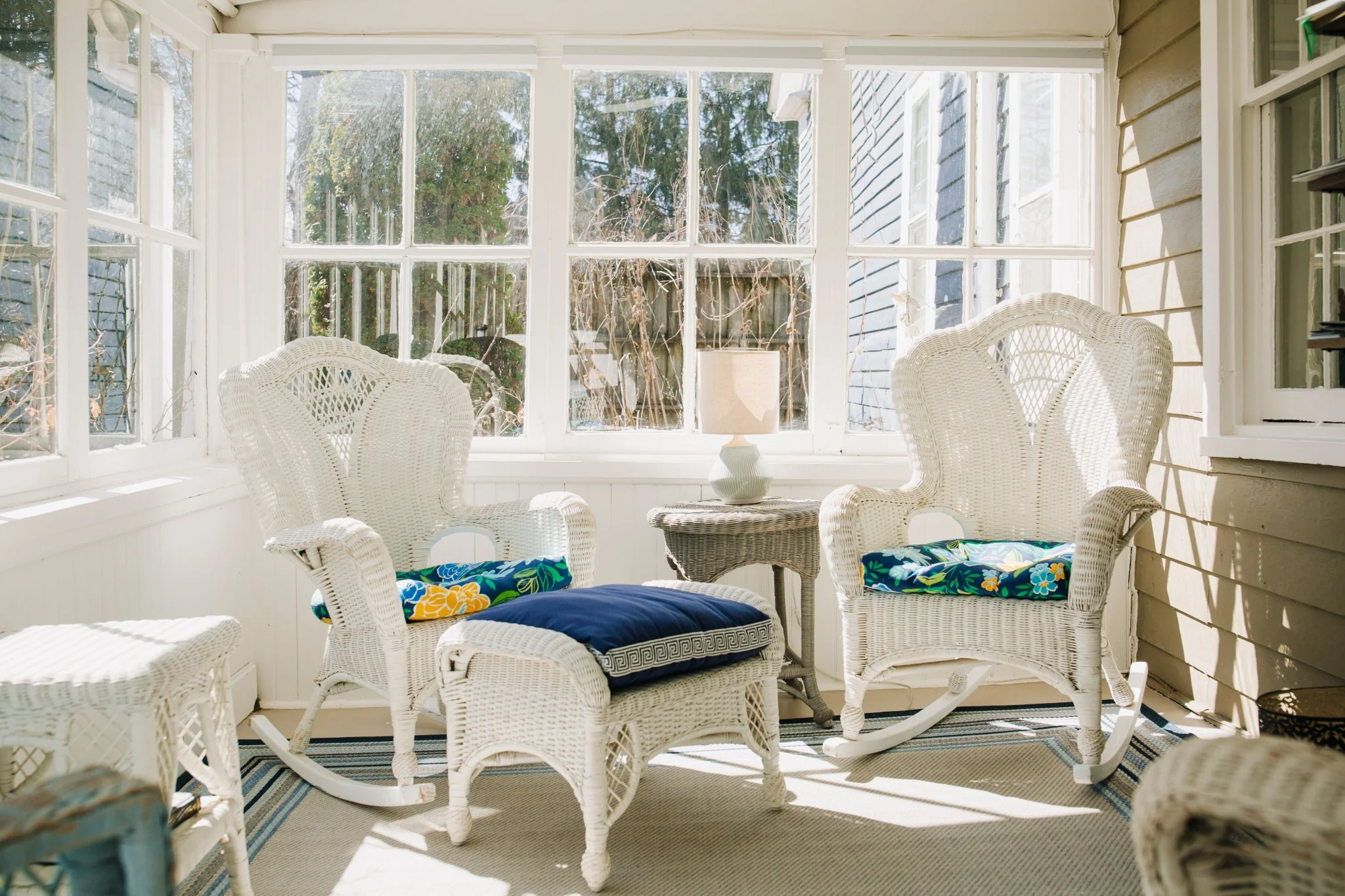 Sunlit porch with white wicker chairs, a small table with a white lamp, and a view of the outdoors through large window panes.