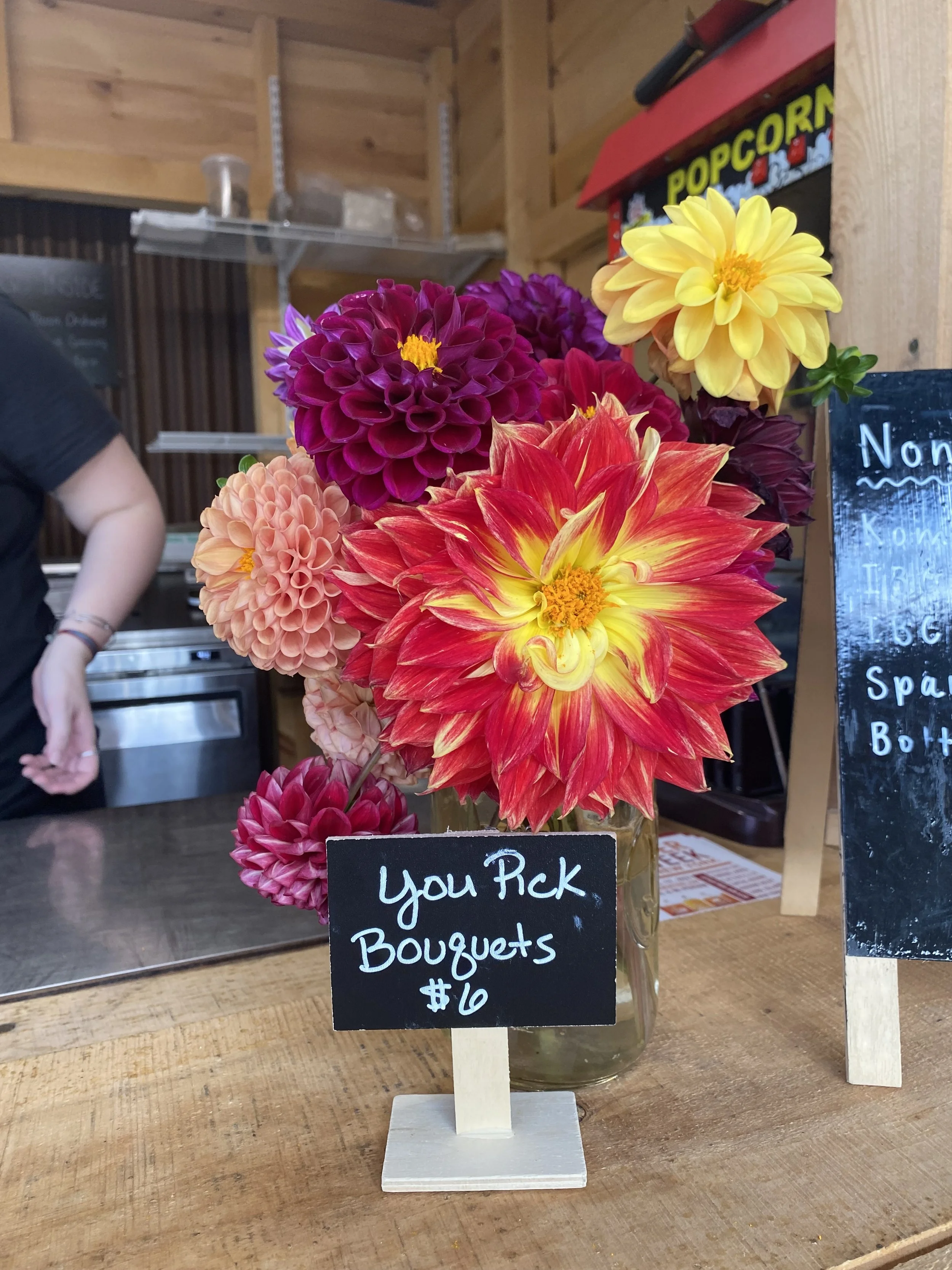 Colorful dahlias arranged in a glass jar on a wooden table with a handwritten sign that reads 'You Pick Bouquets $16' at South Hill Cider.