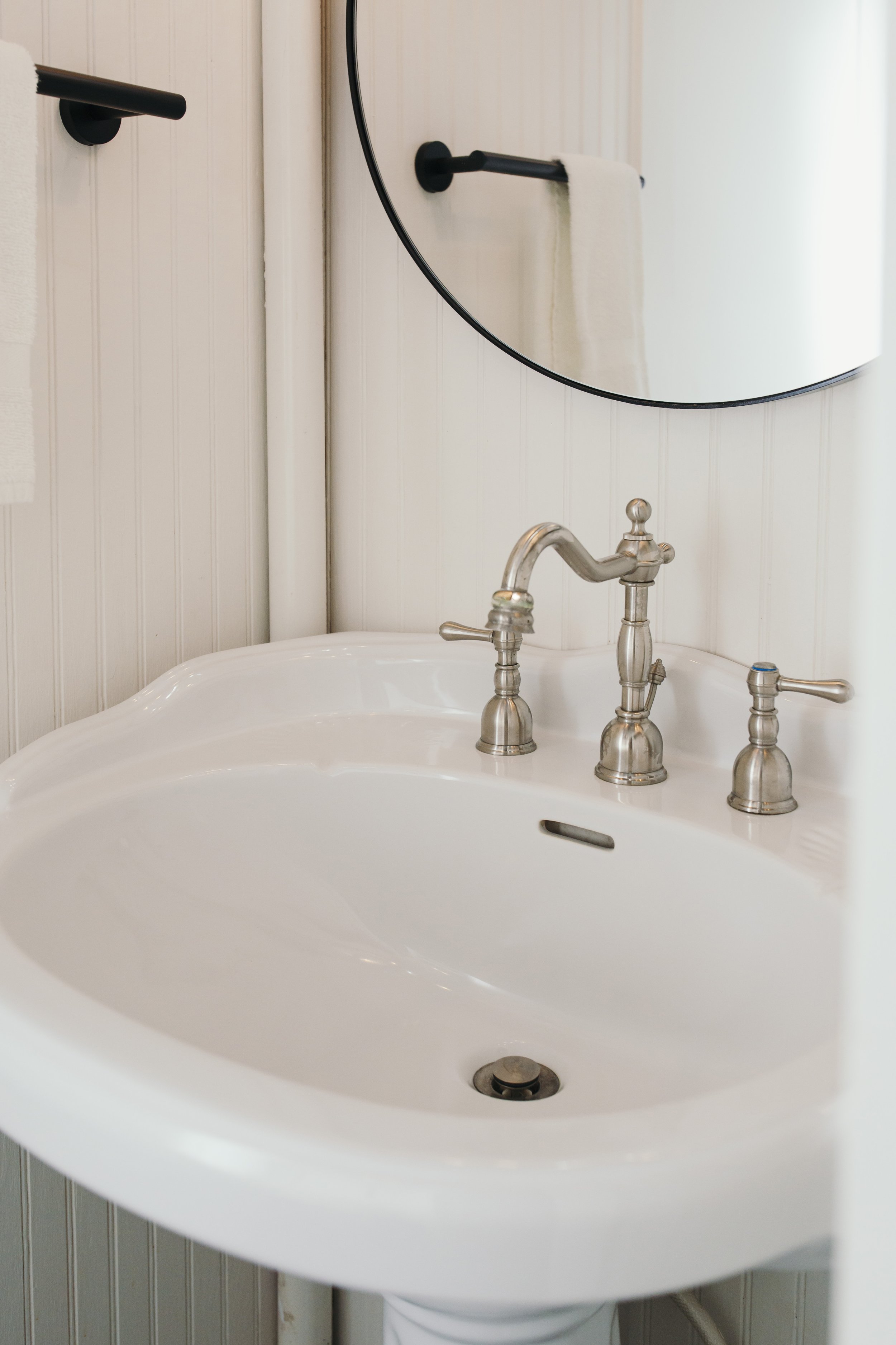 Close-up of a white bathroom sink with a silver faucet and handles, mounted on a wall with light-colored vertical paneling. A round mirror hangs above the sink, partially visible in the image.